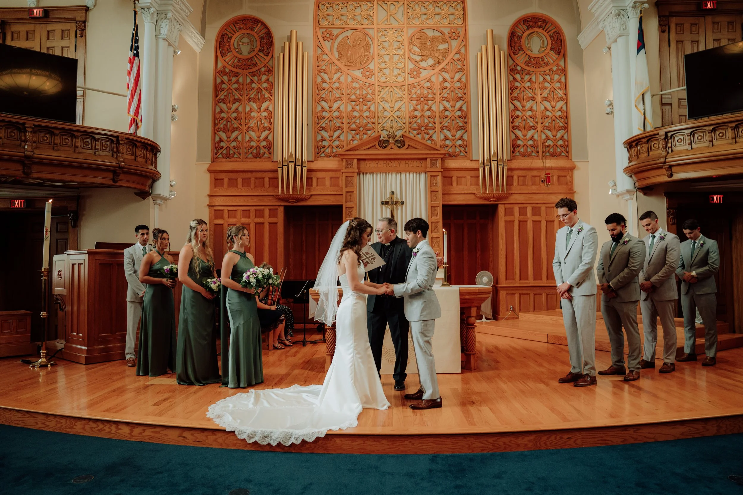 A wedding ceremony inside a church with the bride and groom exchanging vows. Bridesmaids and groomsmen are standing on either side of the couple, with the officiant officiating. The church has wooden accents, religious symbols, and American flags.
