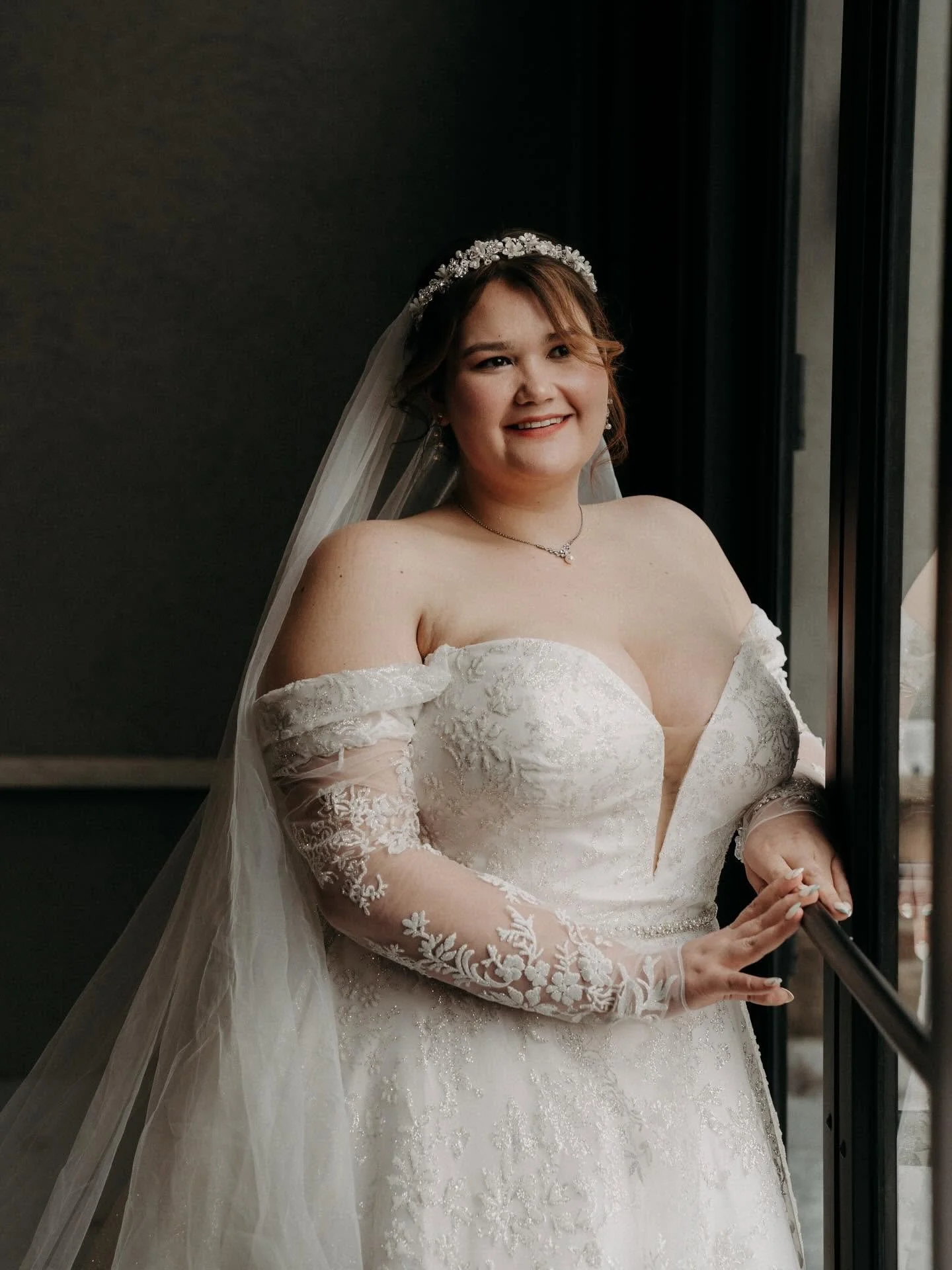 A woman in a wedding dress standing by a window, smiling, with a veil and floral headband, indoors.