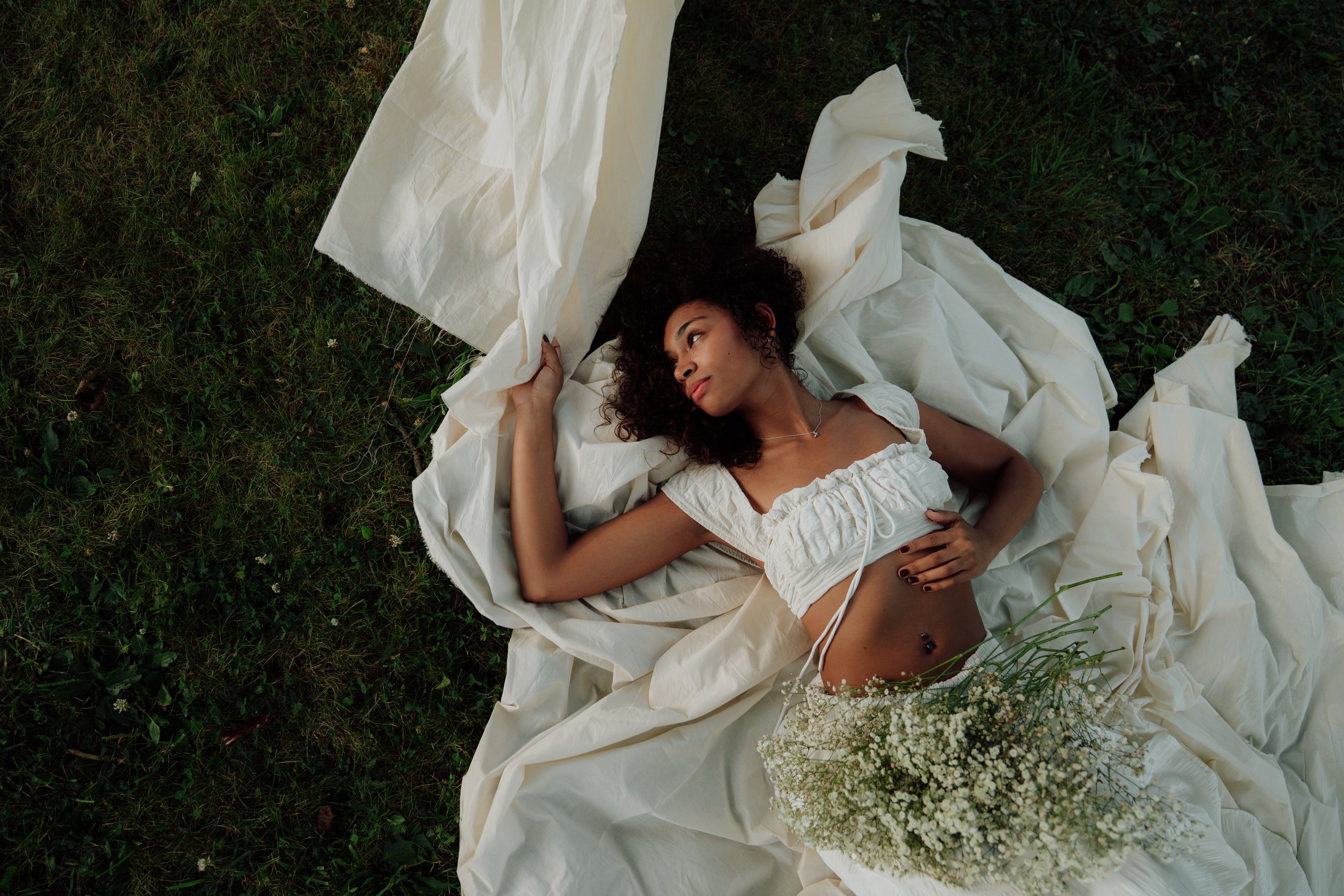 A woman with curly hair lying on grass surrounded by white fabric and flowers, gazing upward.