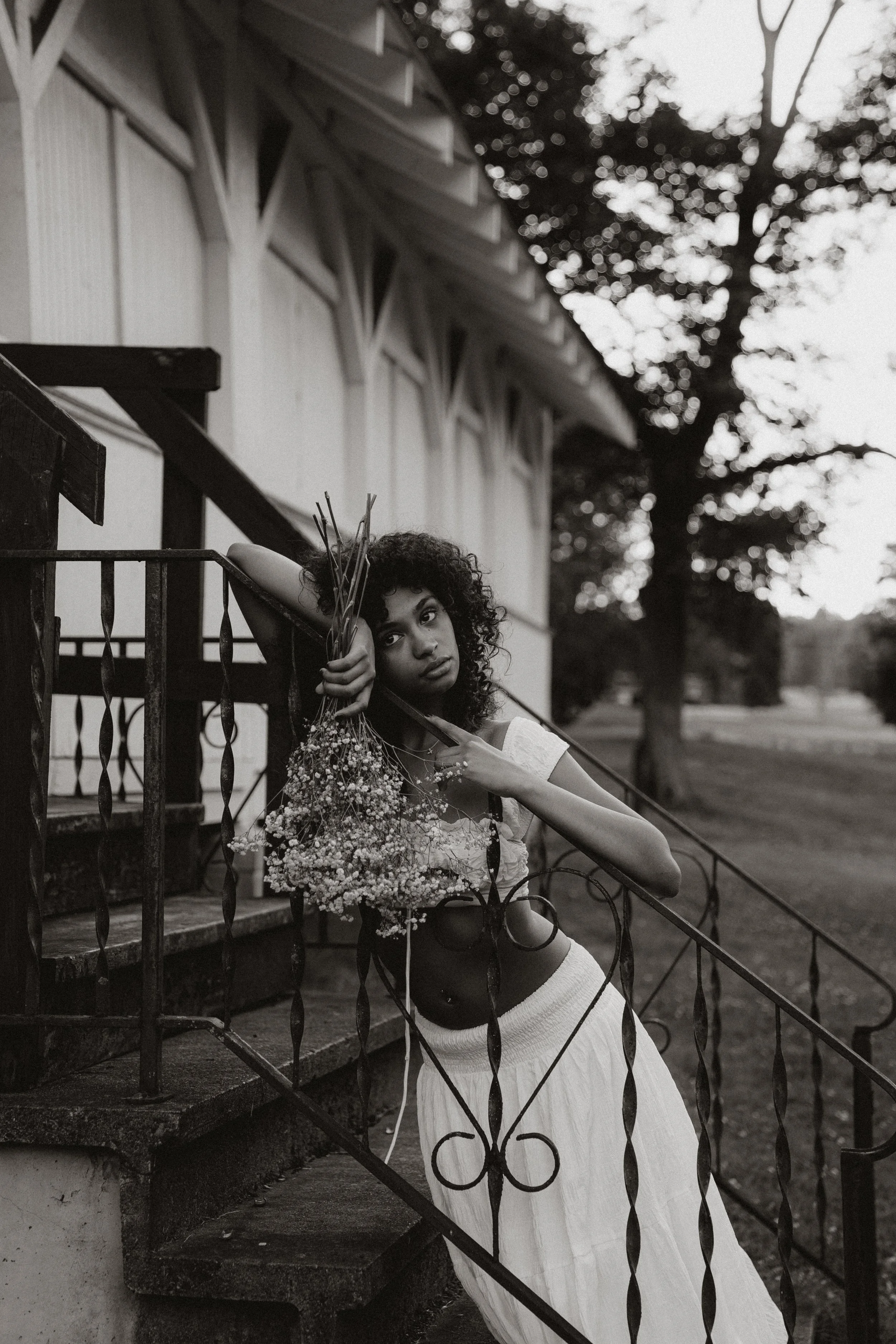 A young woman with curly hair stands on stone steps behind a wrought iron fence, holding a bouquet of flowers. She looks directly at the camera with a neutral expression. There are trees and a building in the background. The photo is in black and whi