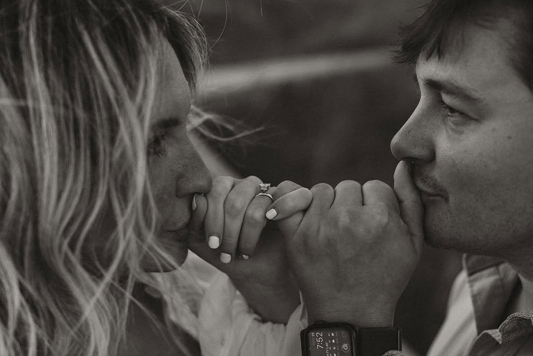 A man and woman with their eyes closed, holding hands with intertwined fingers, gently touching their foreheads and lips in an intimate moment.