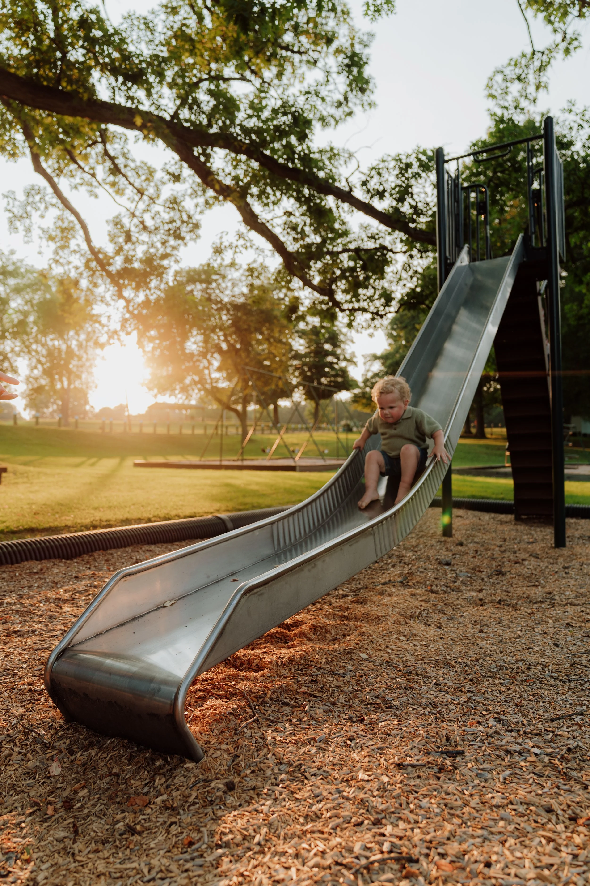 A young boy is sliding down a metal playground slide at a park during sunset. There are large trees with green leaves in the background, and the ground is covered with wood chips.