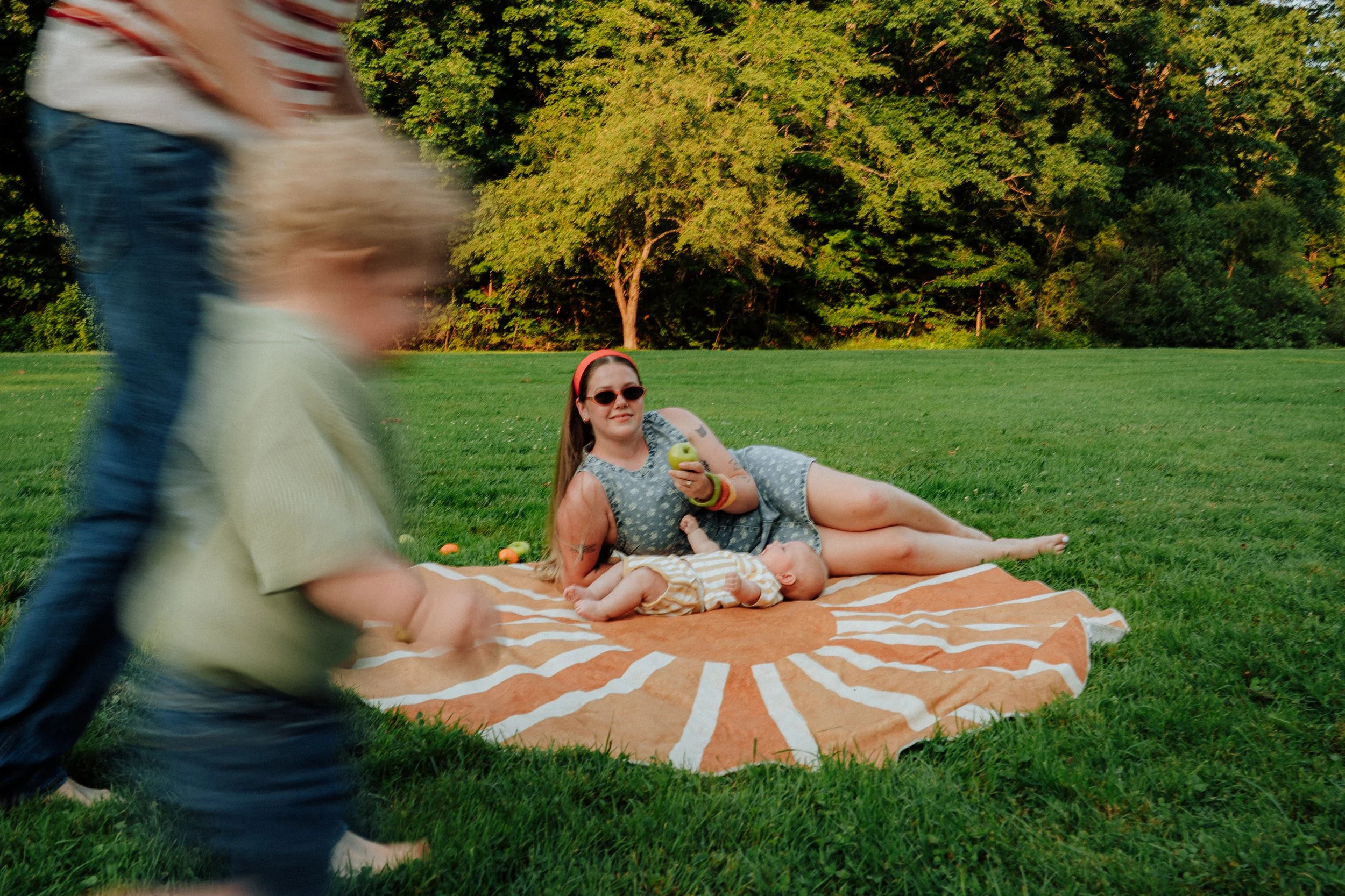 A woman is lying on a striped blanket on the grass, holding an apple, with a small child lying beside her. Two children are walking past in the foreground, out of focus. The scene is outdoors with lush green trees in the background.