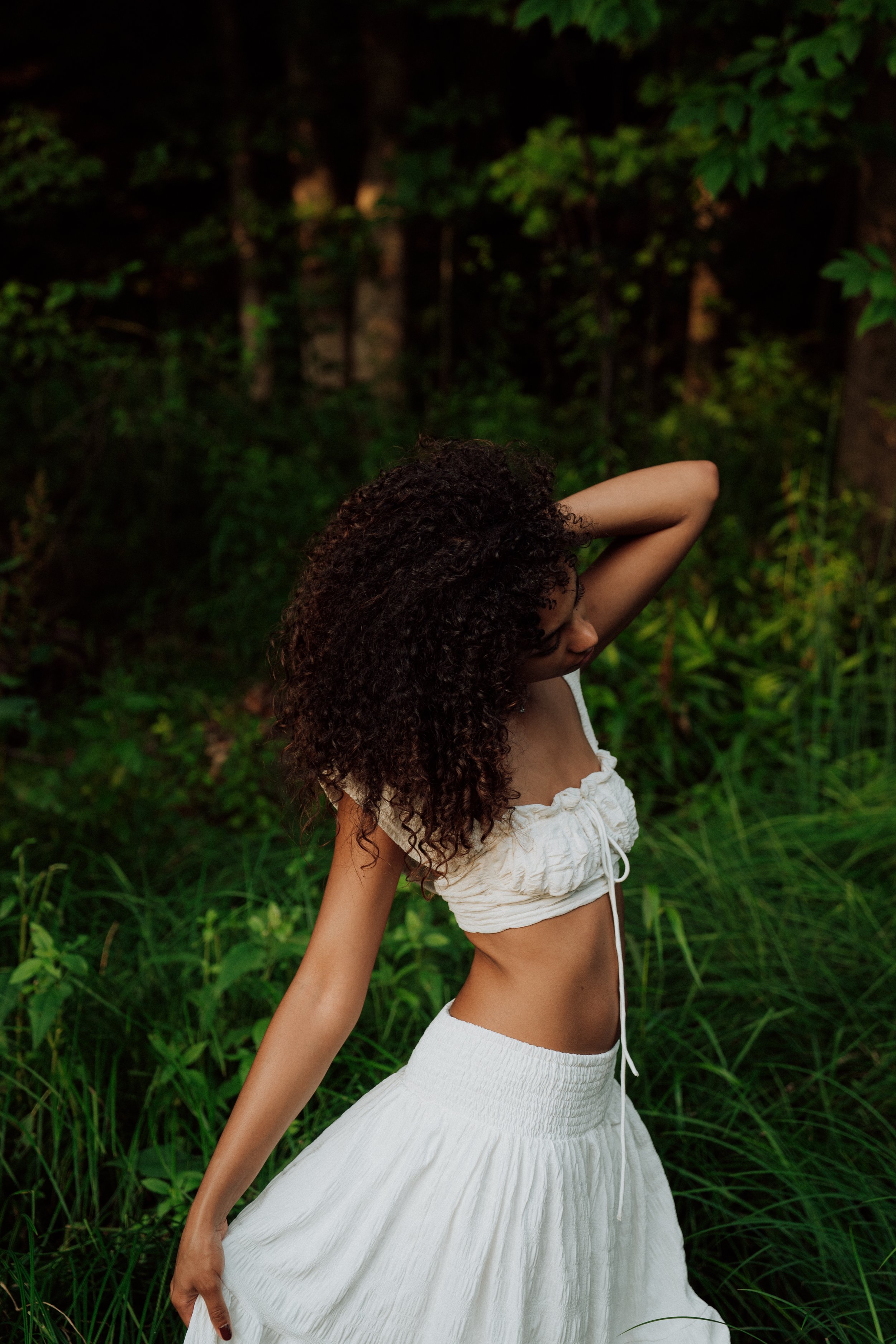 A woman with curly hair standing in a lush green outdoor setting, wearing a white crop top and a white skirt.
