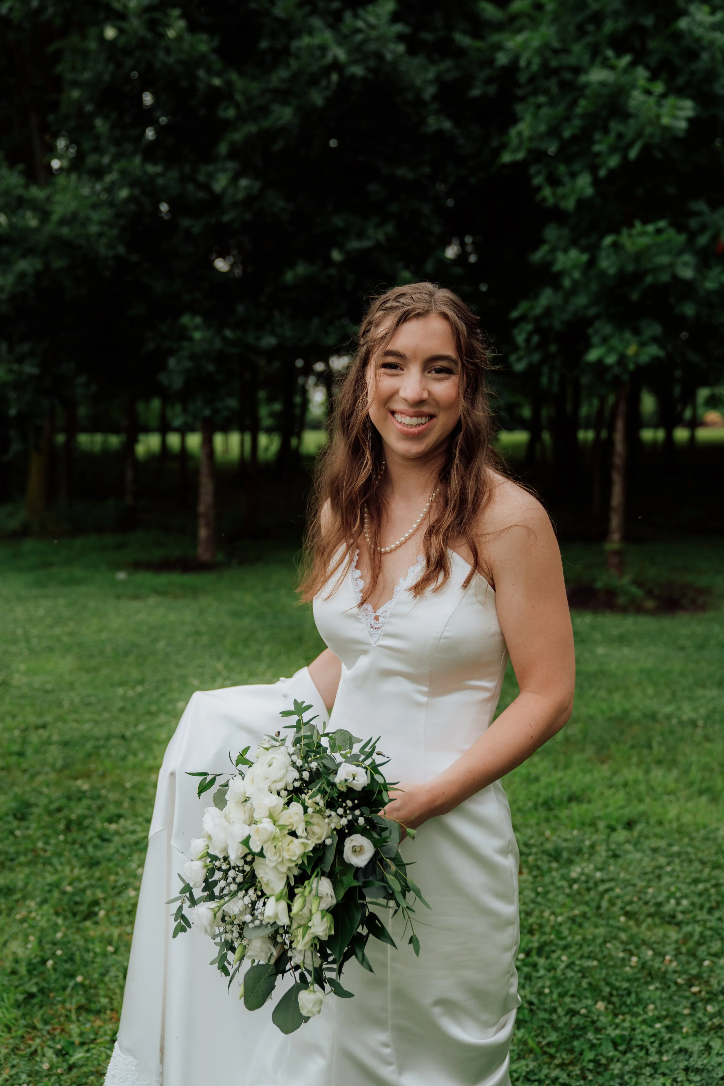 A woman in a white wedding dress holding a bouquet of white flowers and greenery, smiling outdoors with green trees in the background.