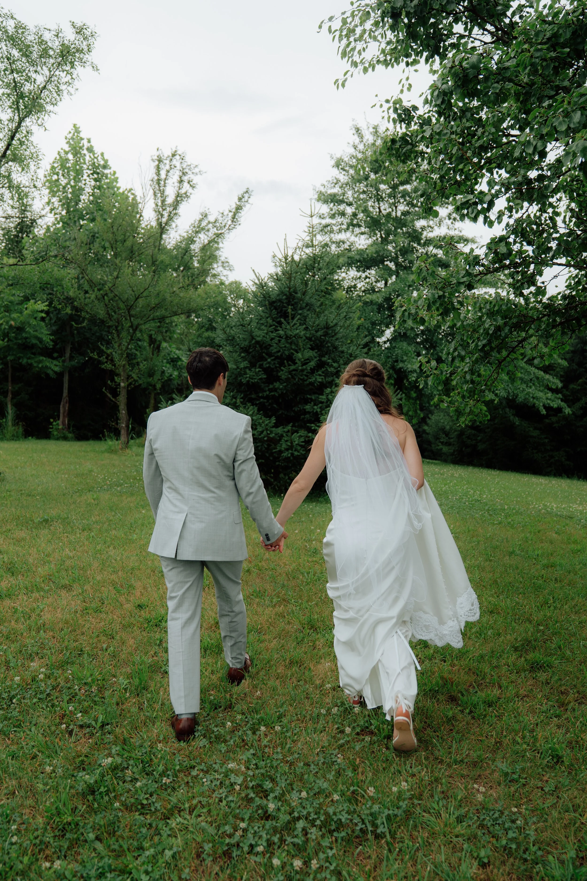 A newly married couple walking hand in hand through a lush green outdoor setting, with trees in the background, during daylight.