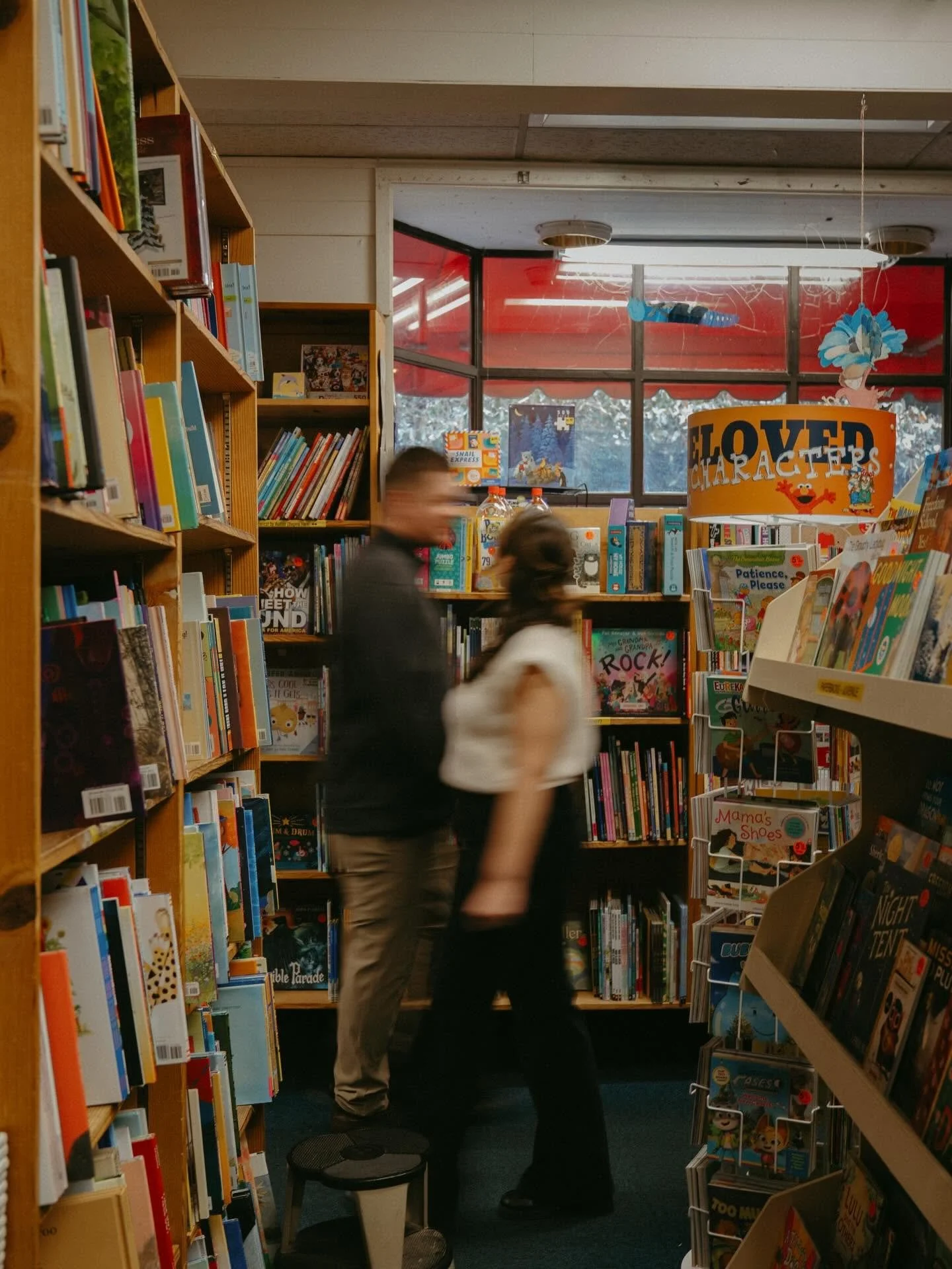 Two people, a man and a woman, standing close together in a bookstore aisle, blurred due to motion, surrounded by shelves of books, with a large window behind them.
