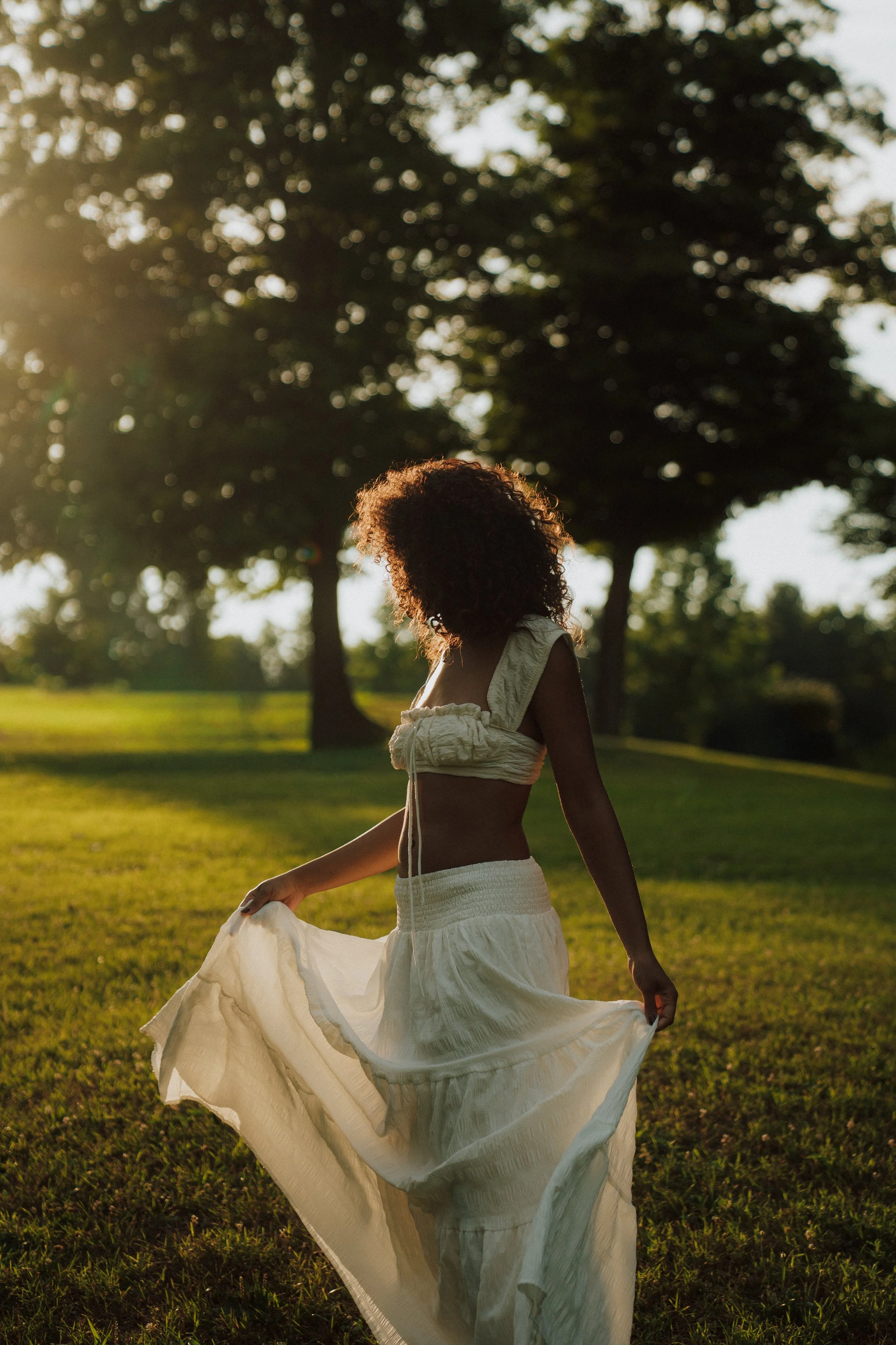 A woman with curly hair wearing a light-colored crop top and flowing skirt, holding the hem of her skirt, walking outdoors on grass during sunset, with trees in the background.