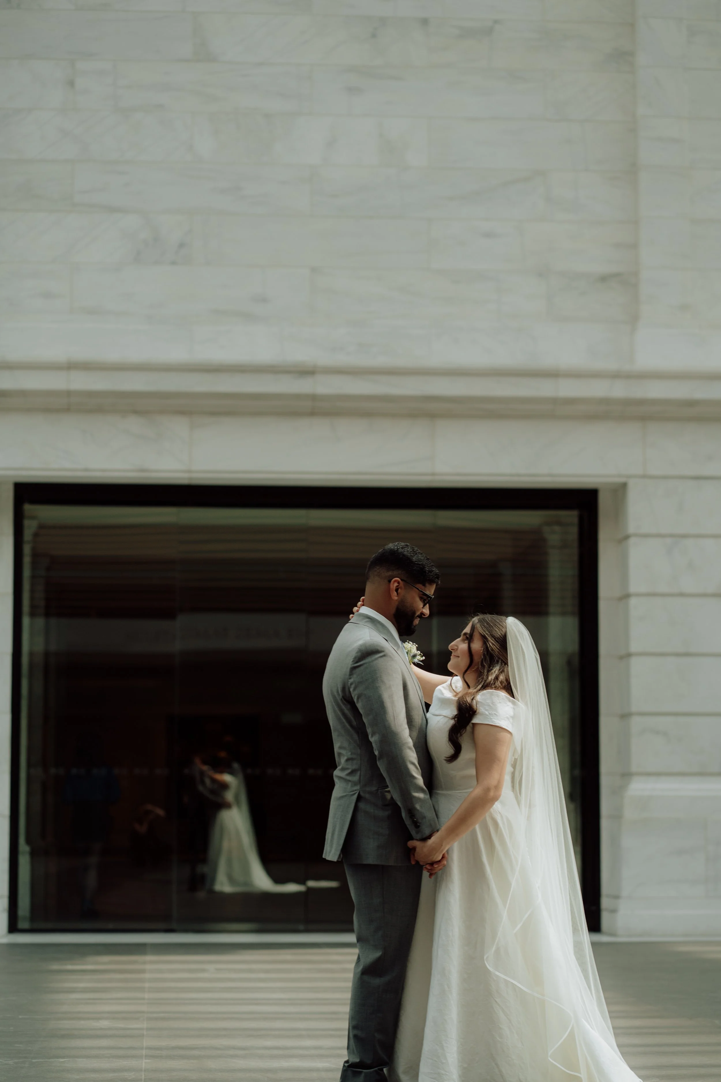 A bride and groom holding hands and standing close together in front of a modern building with large glass windows, smiling at each other.
