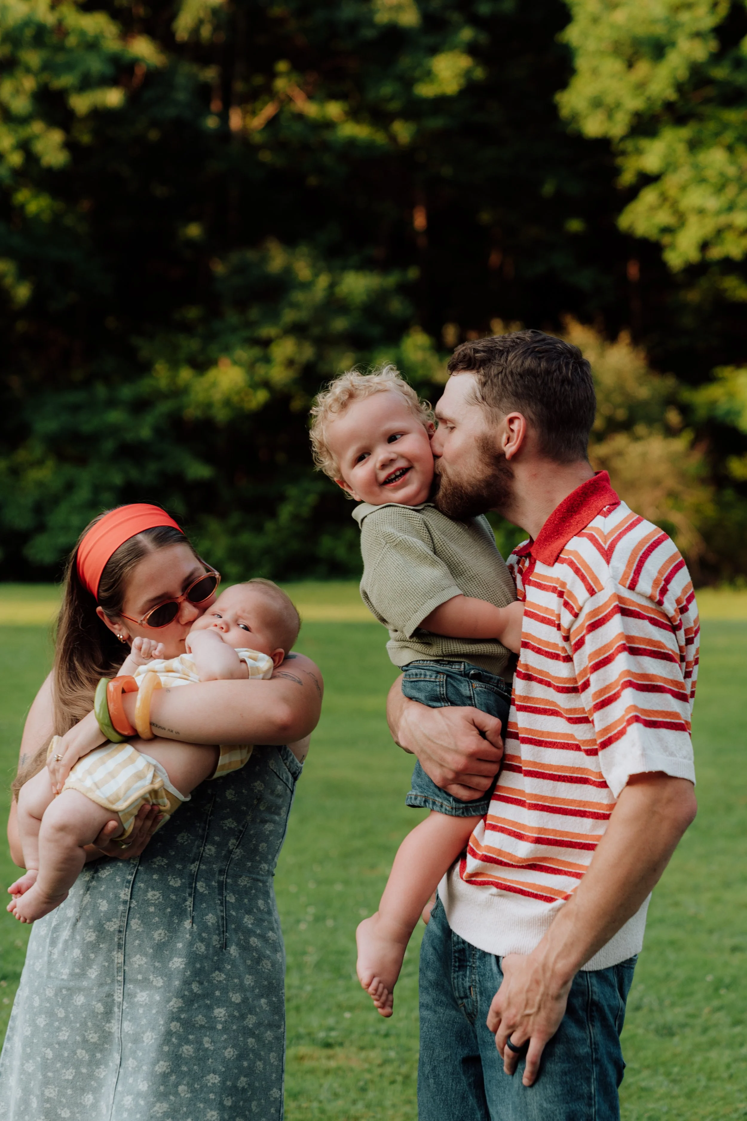 A family outdoors on a grassy field with trees in the background, consisting of a woman holding a baby, a man kissing a young boy, and another child partially visible.