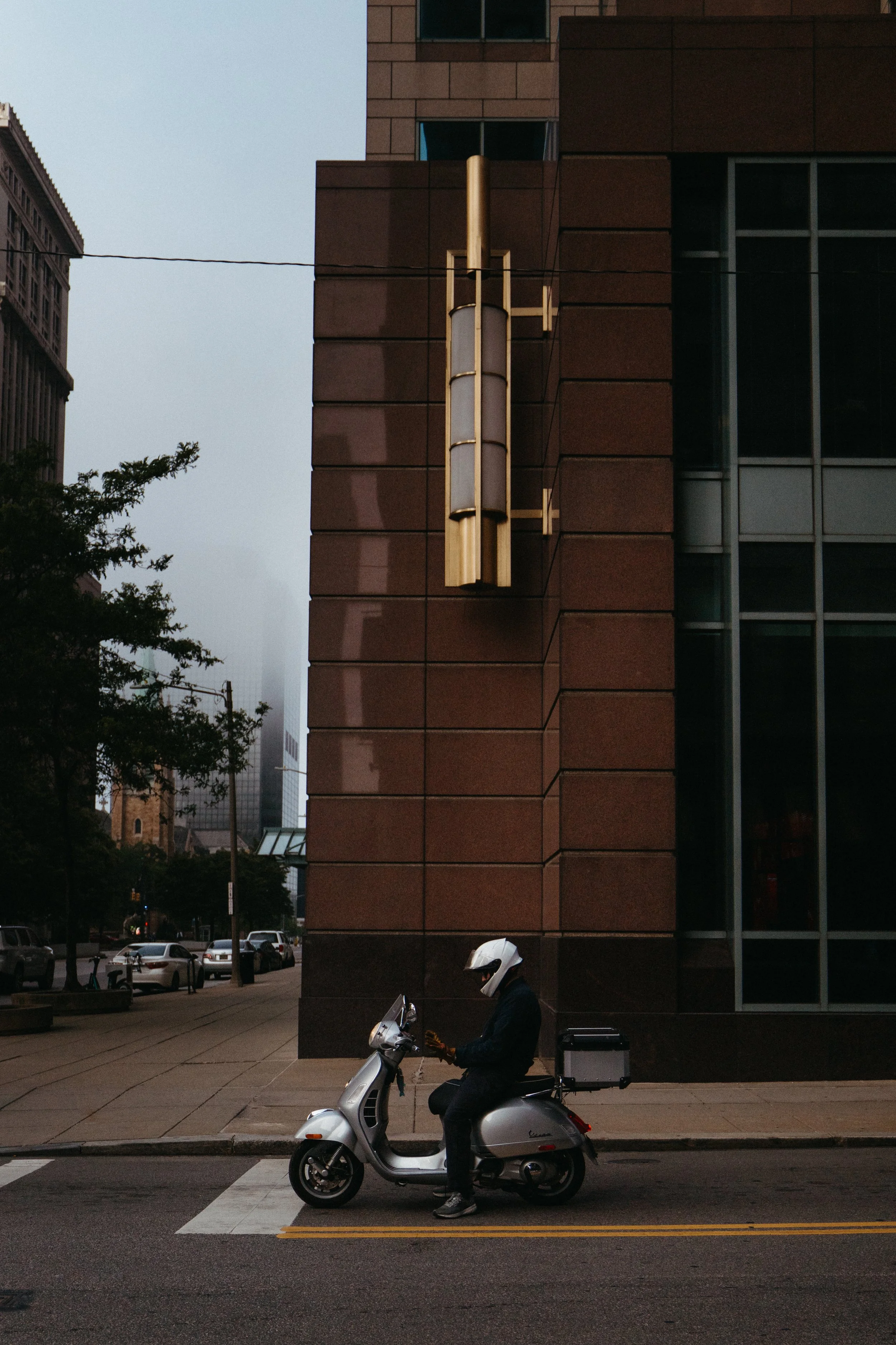 A person wearing a helmet riding a silver scooter on a city street, with a large dark building and trees in the background.