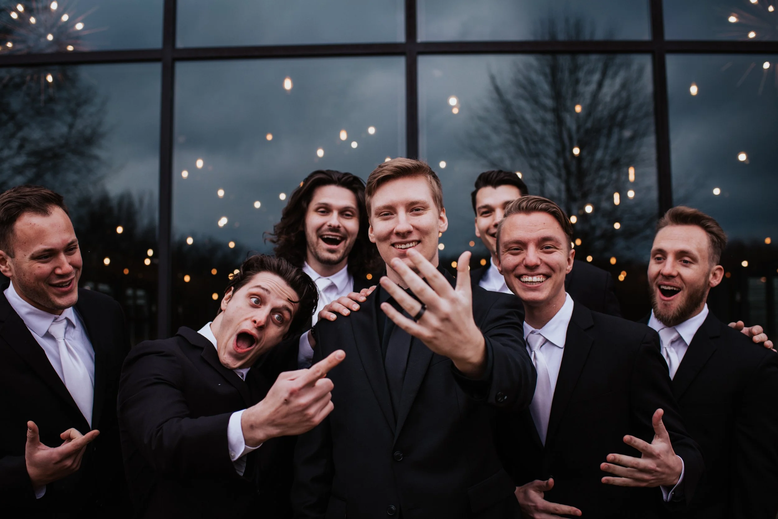 A group of six men dressed in formal suits and ties taking a selfie outside in front of a large window with string lights and trees reflected on it, during overcast weather.