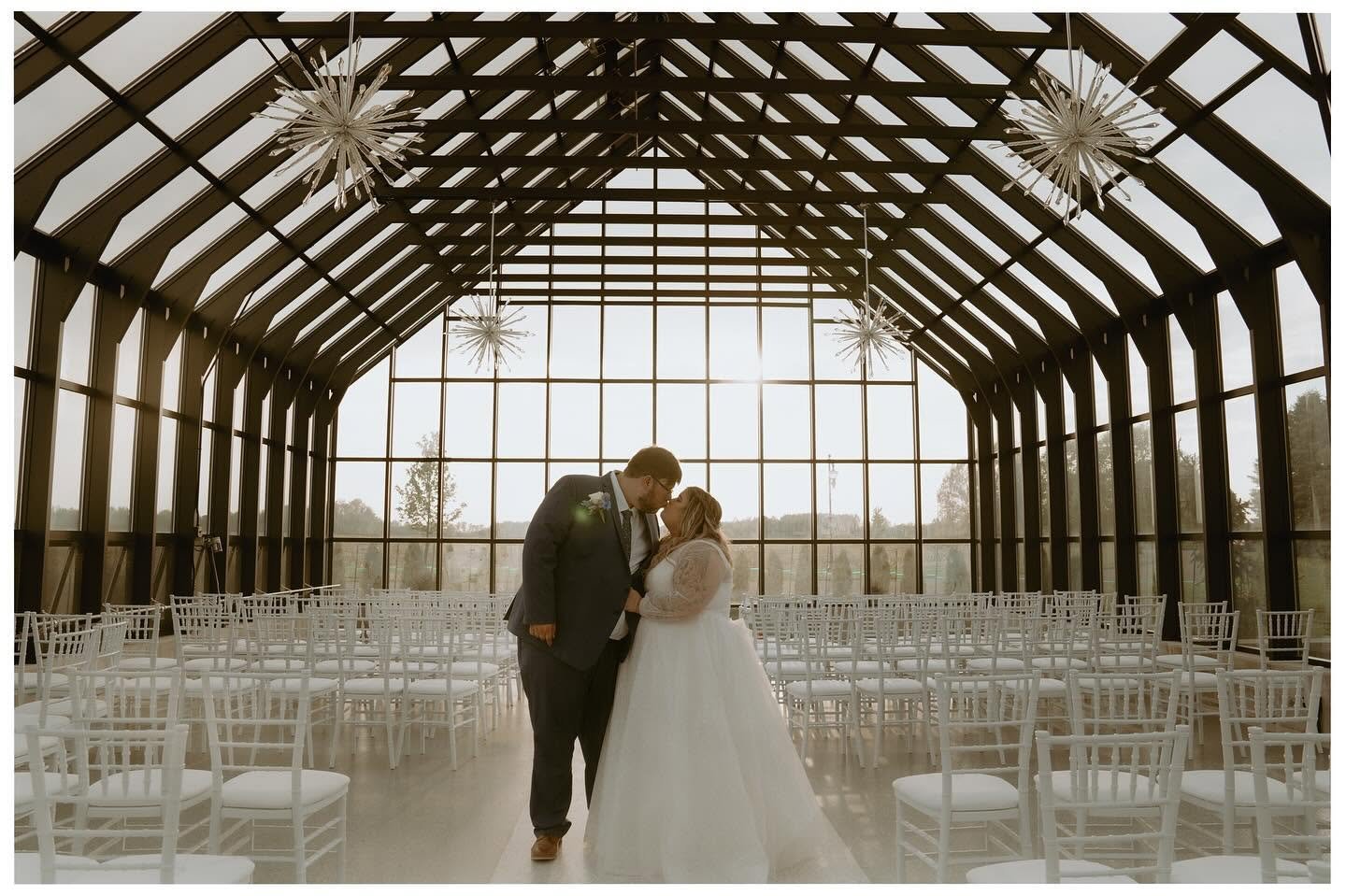A newlywed couple shares a moment inside a glass chapel with a high arched ceiling, surrounded by empty white chairs, sunlight streaming through large windows.