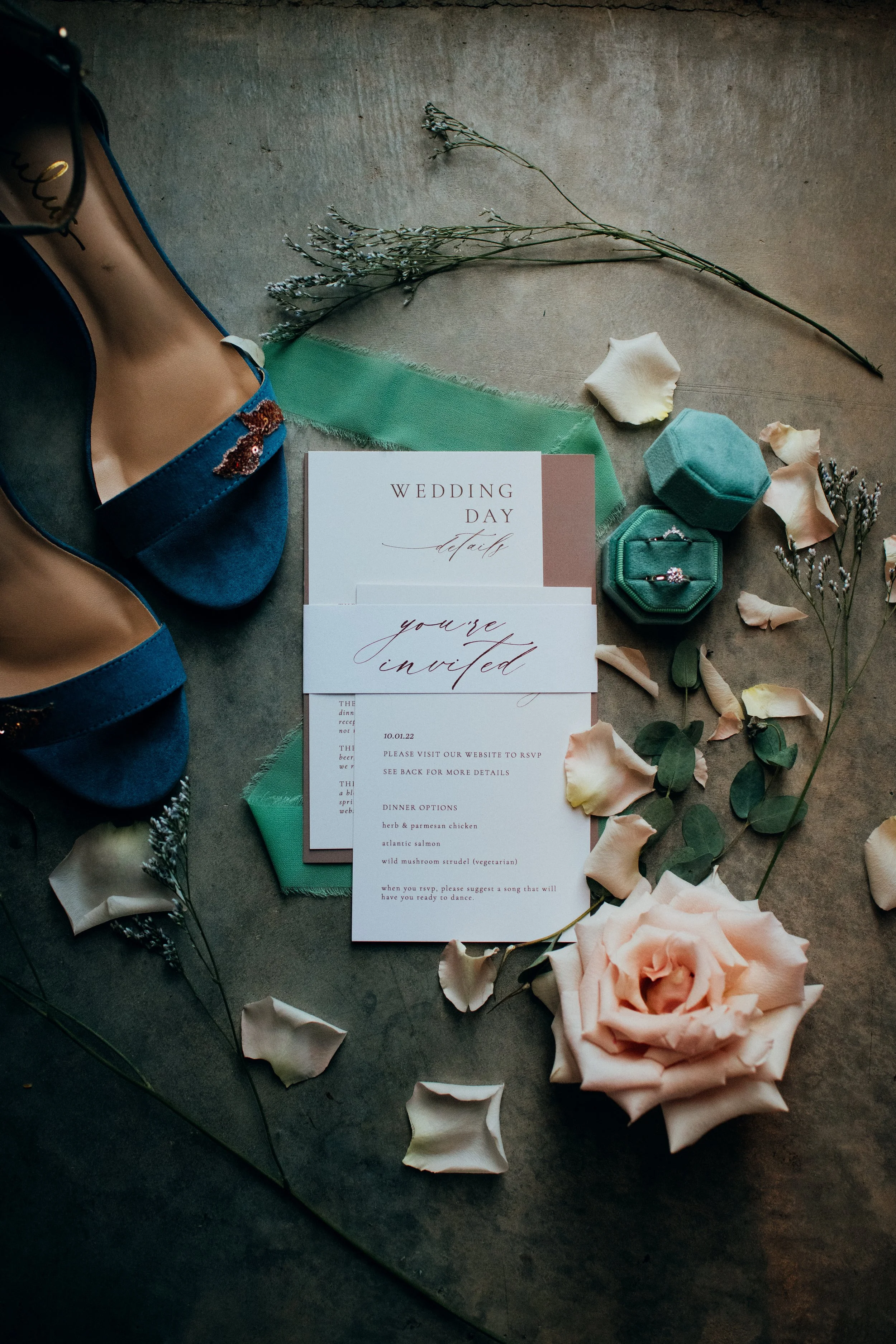 Wedding invitation cards, dried flowers, rose petals, a green fabric, a pair of rings in a green ring box, a pair of blue shoes, and a large pink rose flower on a gray surface.