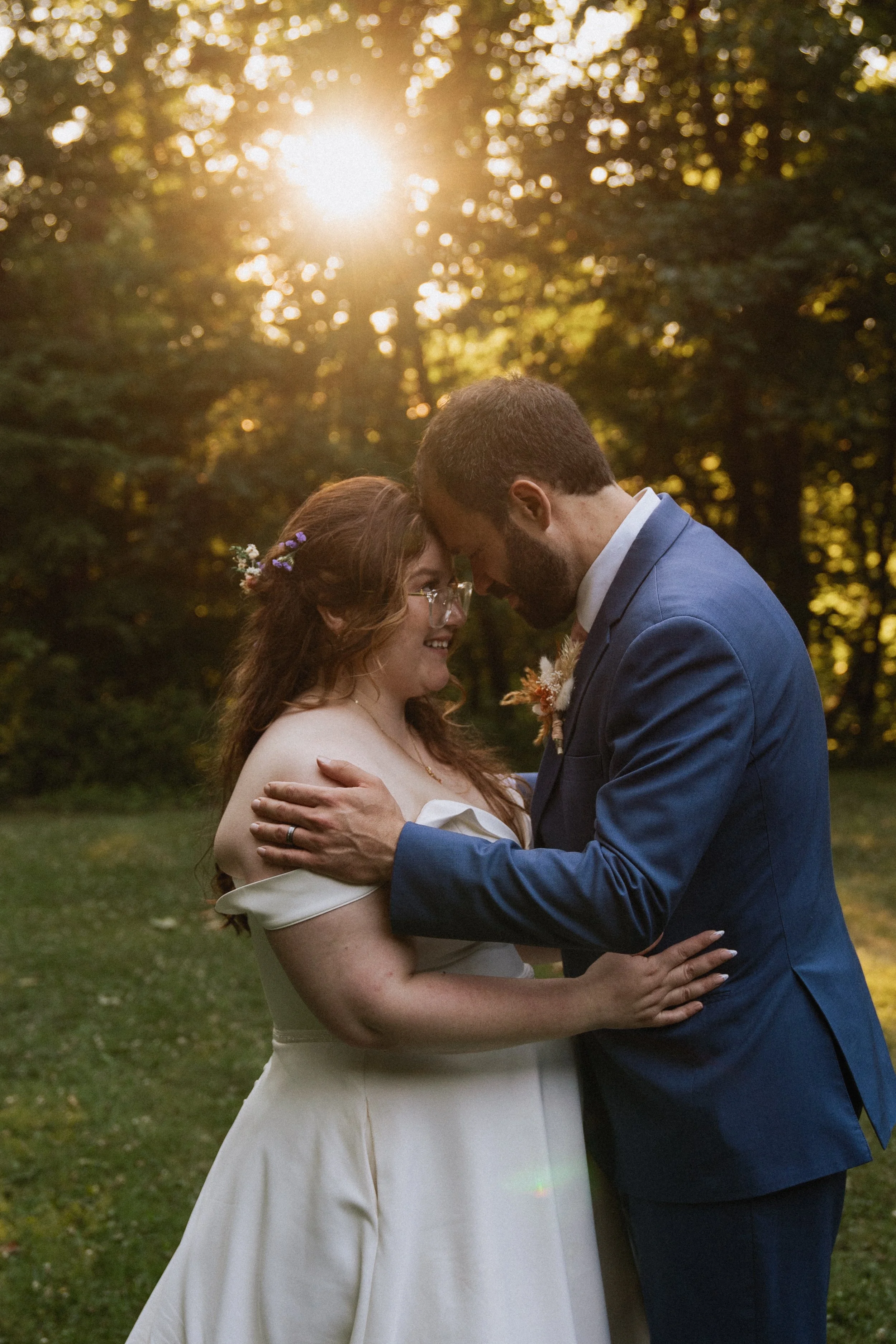 A couple sharing a romantic moment in a park during sunset, the woman with long wavy hair and glasses, wearing a white dress, and the man in a blue suit, leaning their foreheads together.