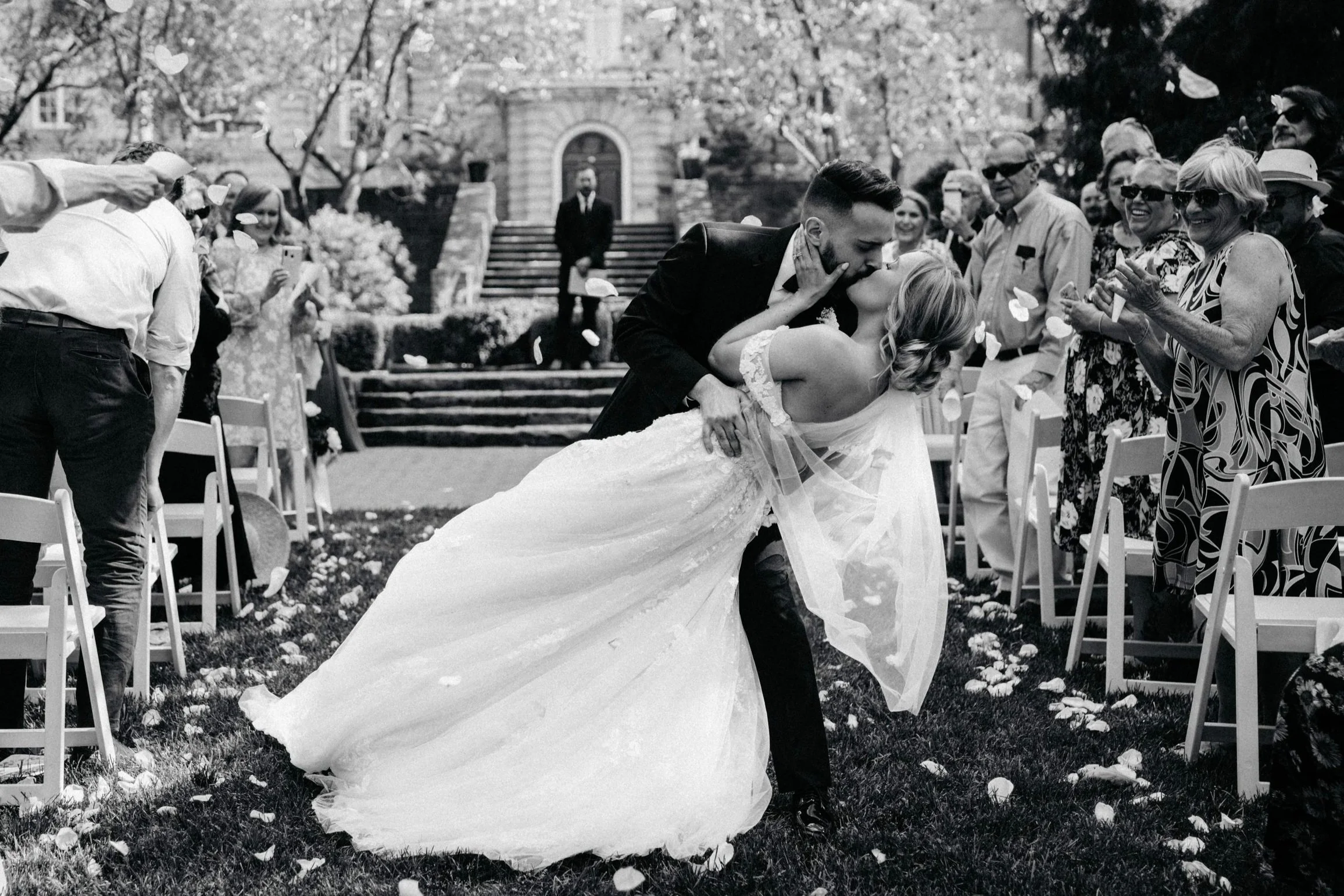 A bride and groom sharing a kiss during their outdoor wedding ceremony, surrounded by guests seated on either side, with flower petals scattered on the ground and trees in the background.