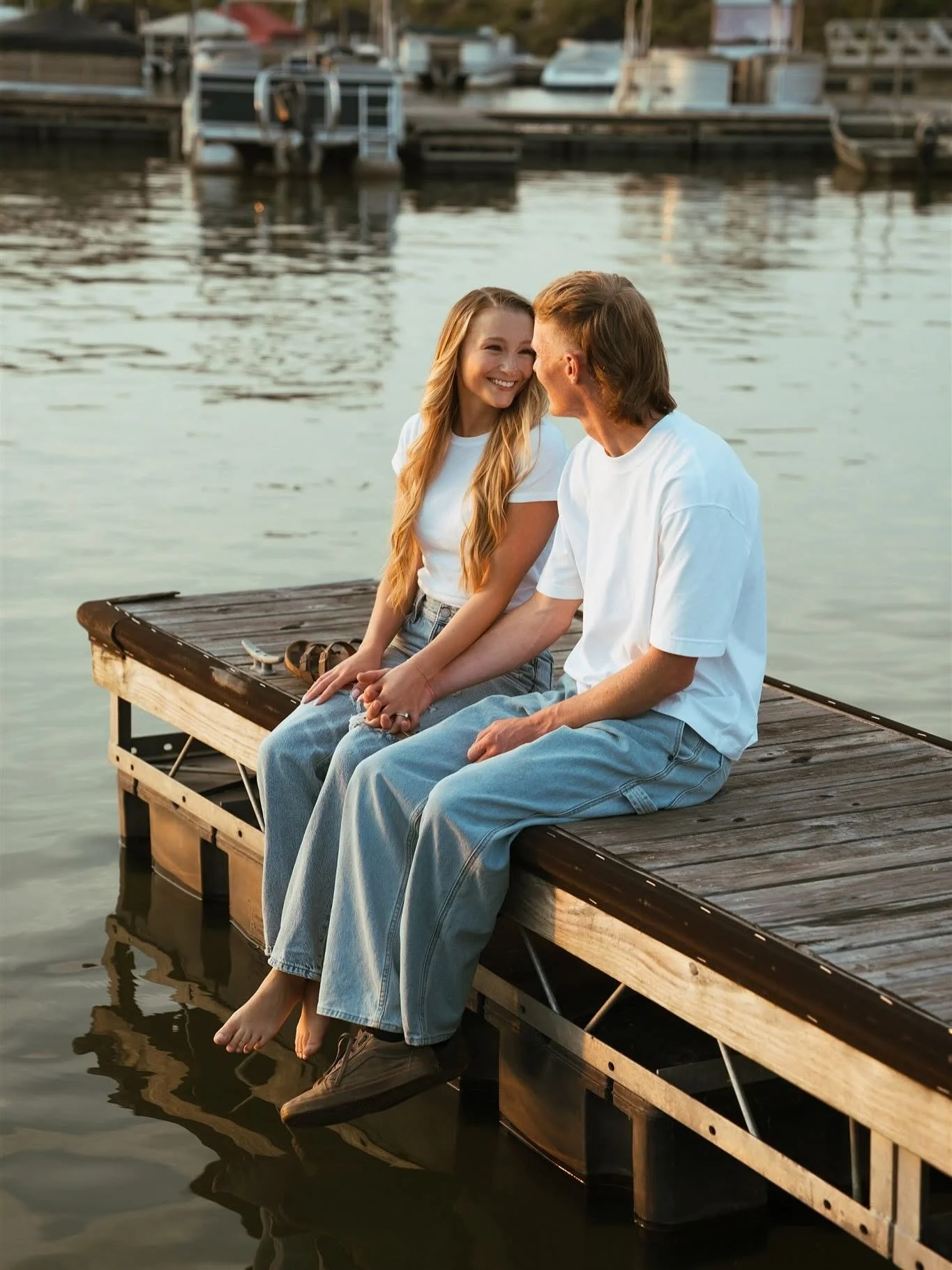 A young woman and a young man sitting on a wooden dock by the water, smiling at each other, with boats and a marina in the background.