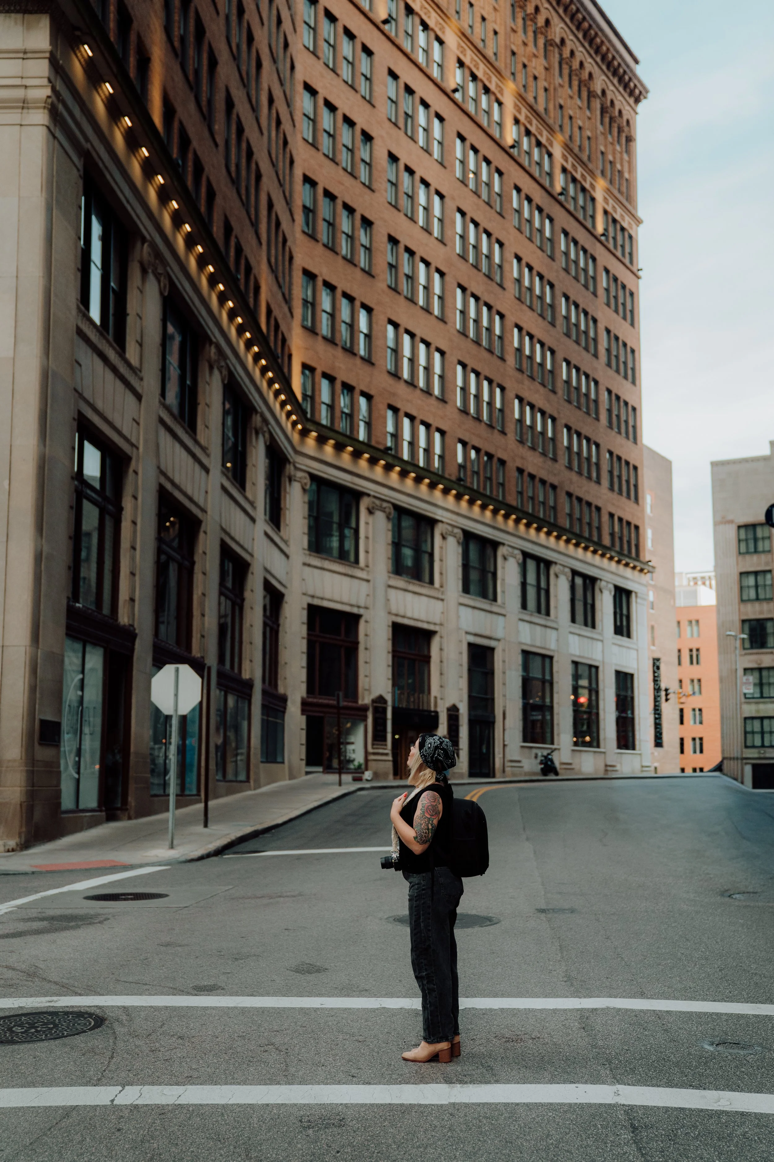 A woman with tattoos on her arms, wearing a beanie, glasses, a black shirt, and jeans, standing on a city street corner with tall buildings in the background. She has a backpack and a camera hanging around her neck.
