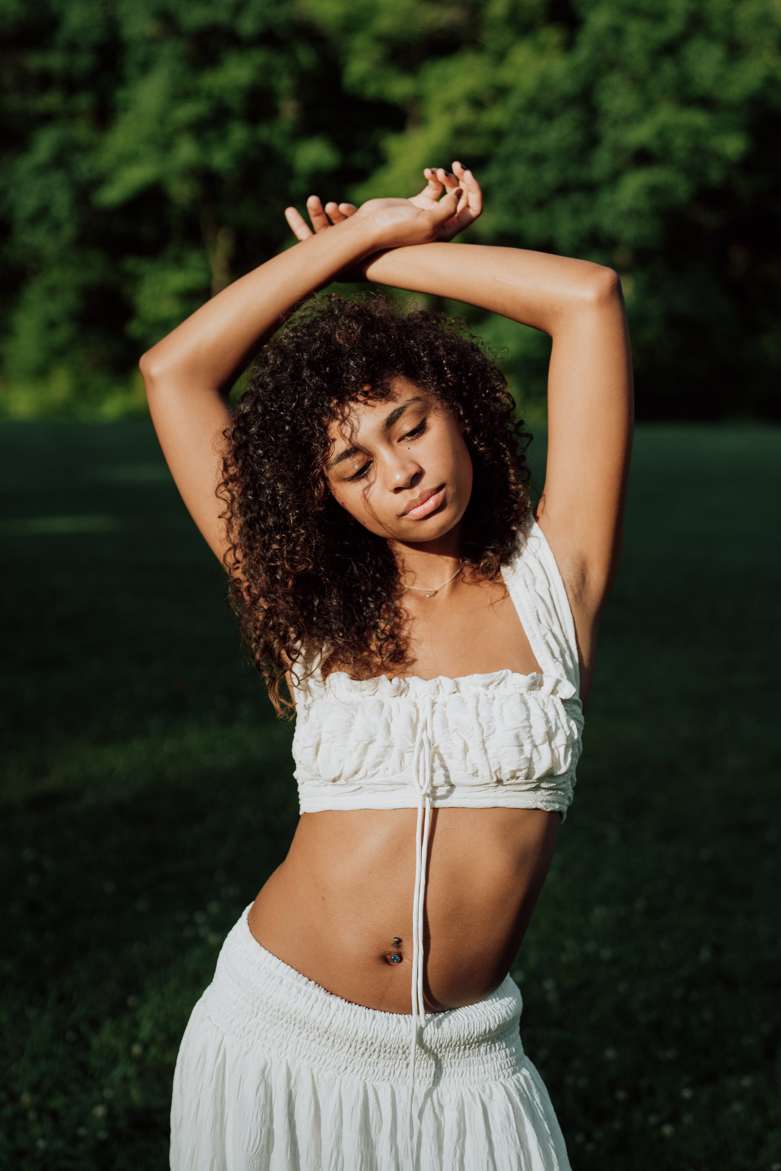 A woman with curly hair outdoors during daytime, wearing a white crop top and skirt, with her arms raised above her head and eyes closed.