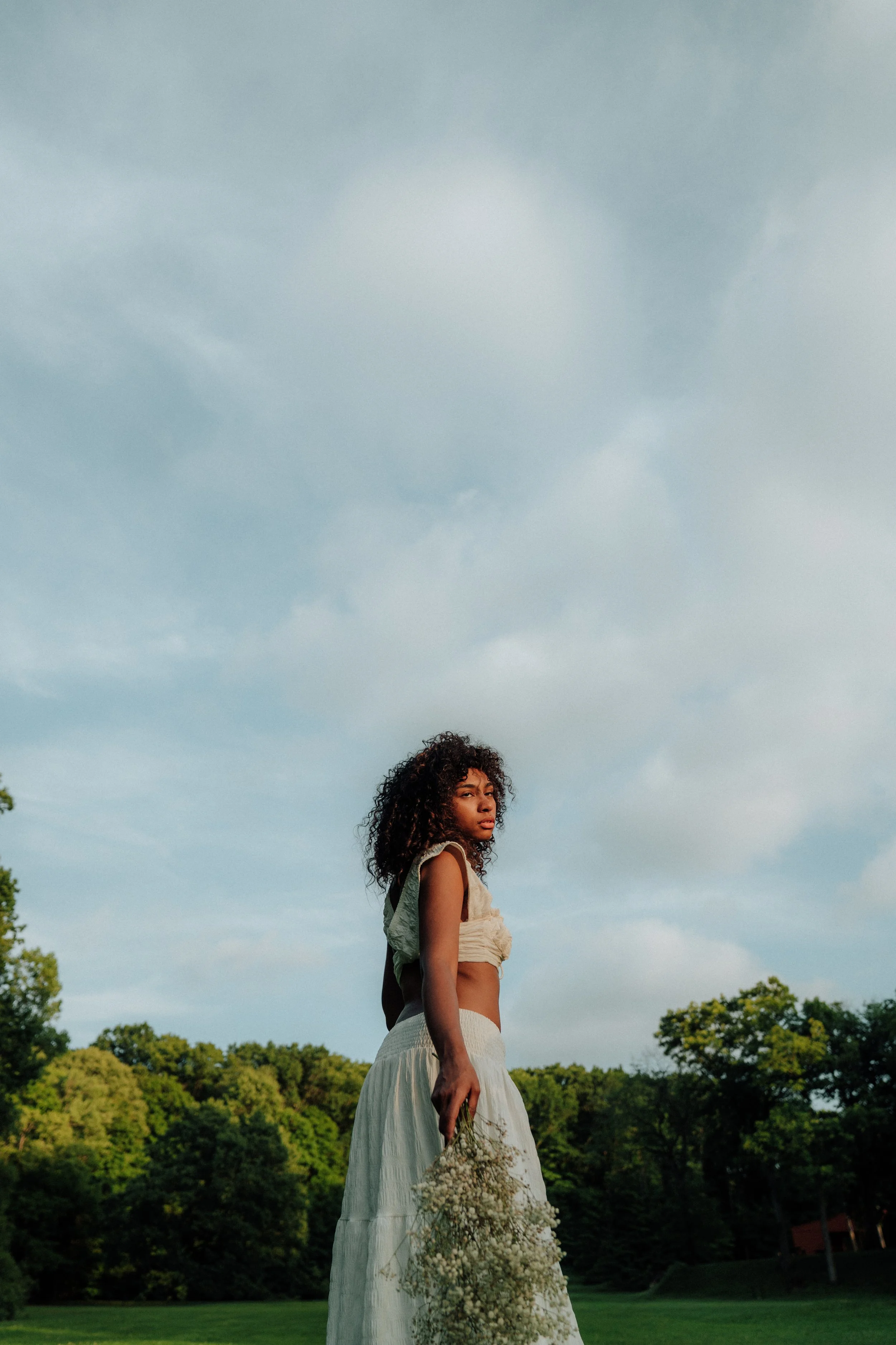 Young woman with curly hair standing outdoors in a dress, holding a bouquet of flowers, with trees and a cloudy sky in the background.