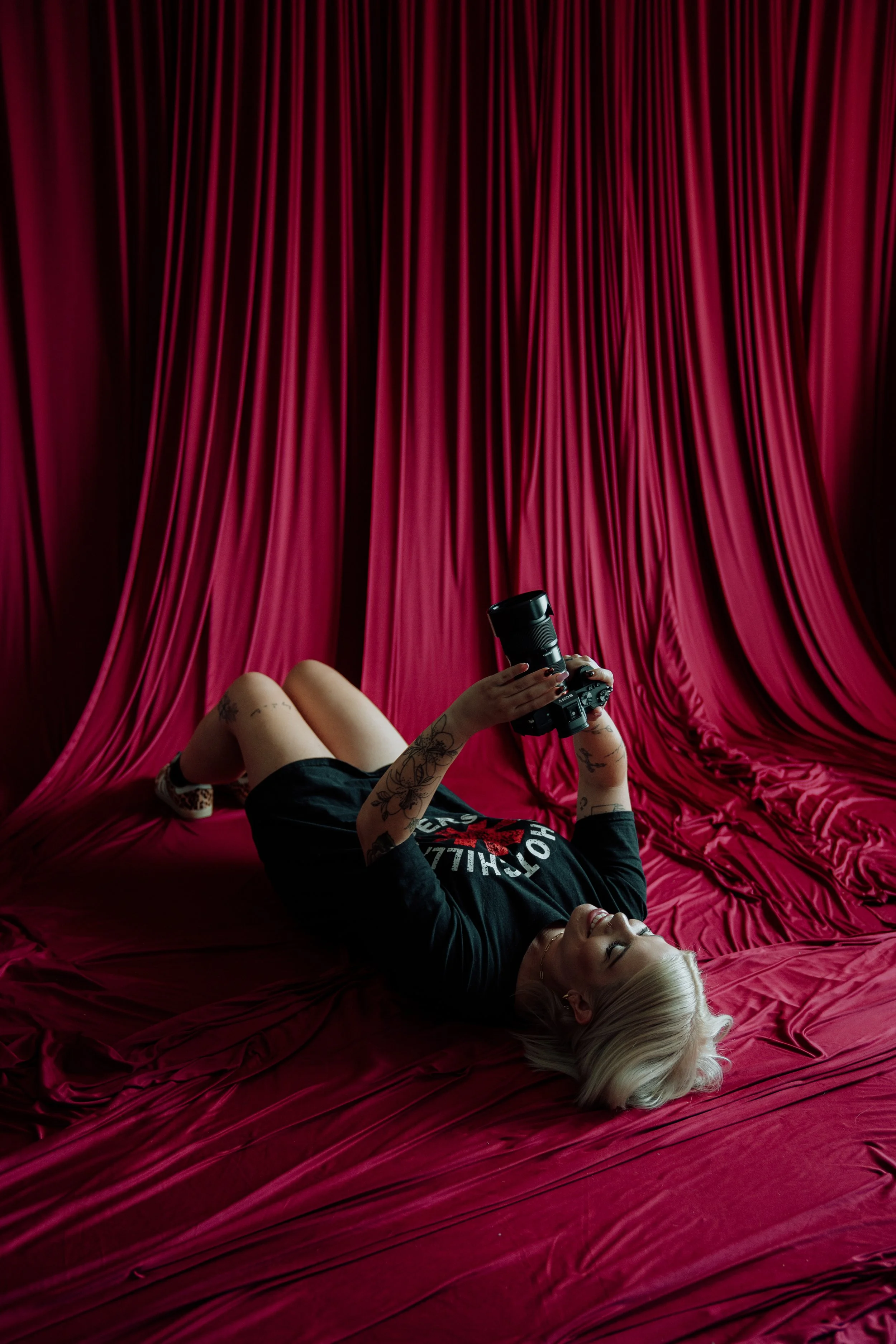 A woman with blonde hair and tattoos is lying on a red satin bed, holding a camera above her. She is wearing a black sweatshirt.