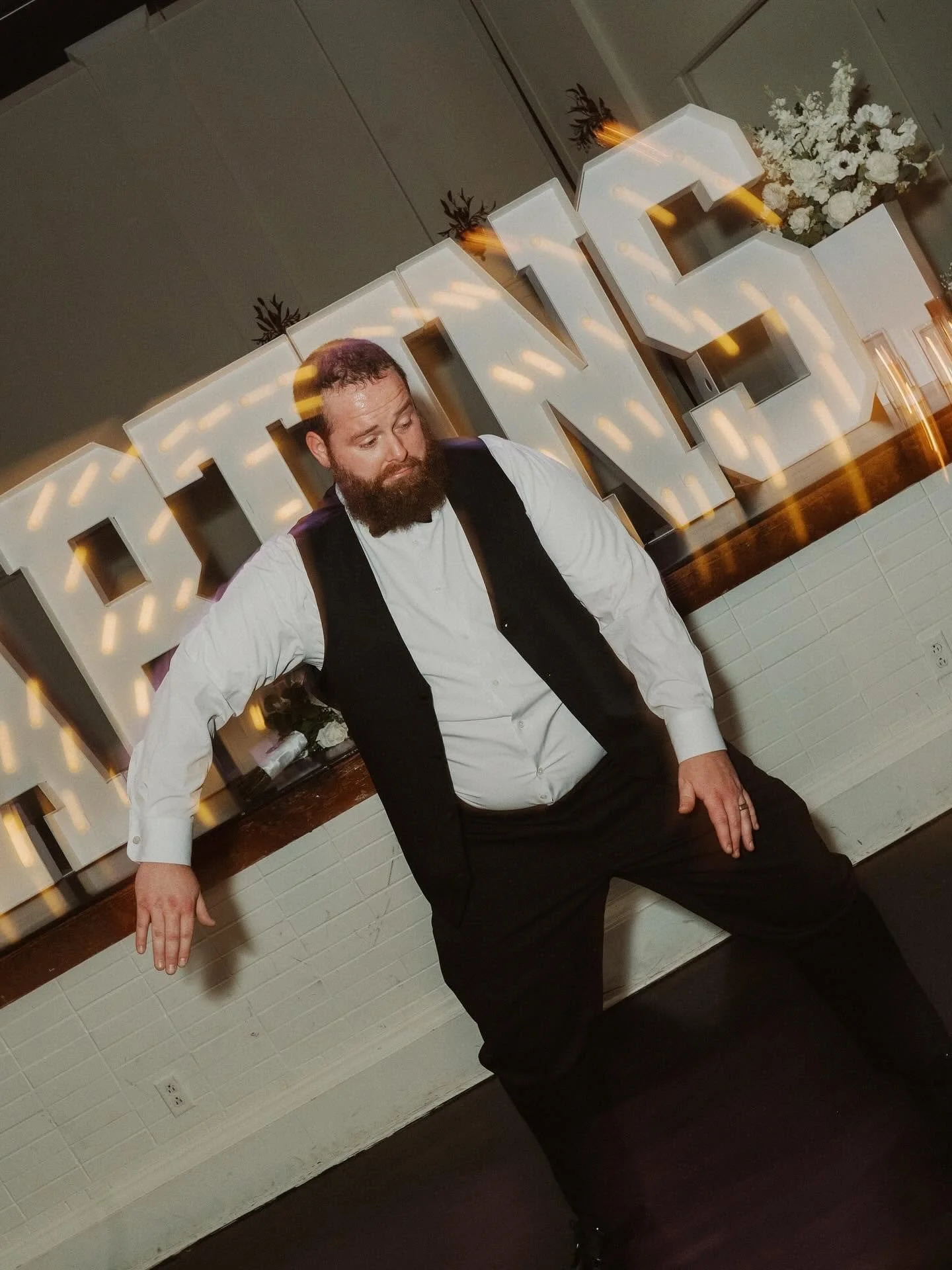 A man with a beard and curly hair wearing formal attire, leaning against a wall with large illuminated letters behind him that spell out 'LOVE'. There are white flowers and potted plants in the background.