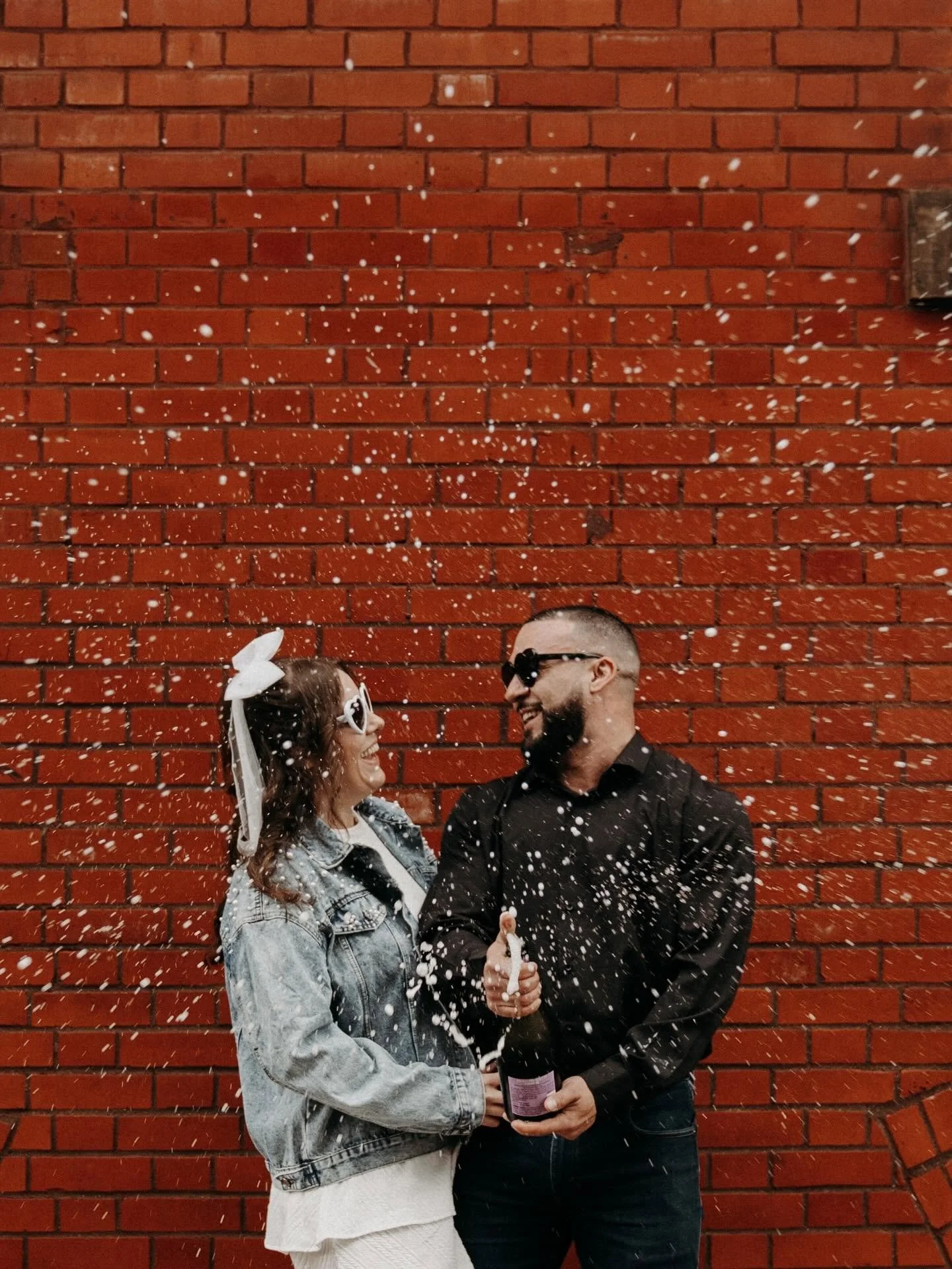 A smiling woman and man stand in front of a red brick wall, holding a champagne bottle and a champagne flute. They are looking at each other, and white confetti is falling around them.
