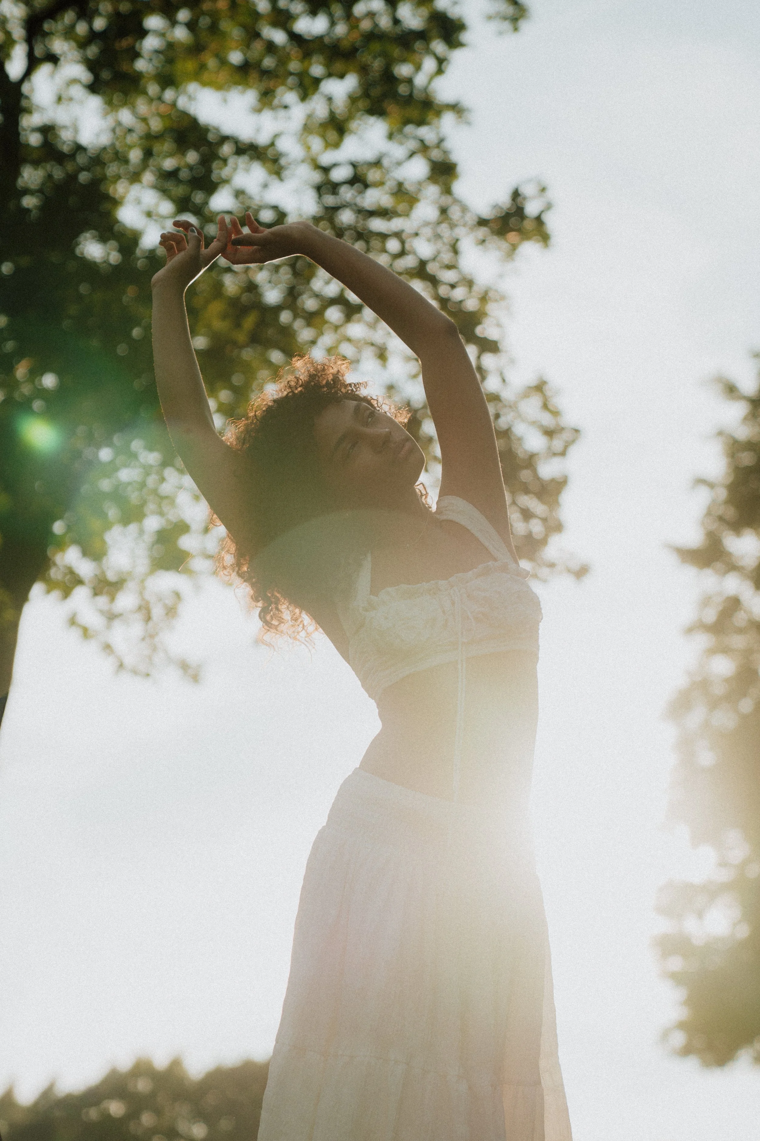 A woman with curly hair wearing a light-colored dress, standing outdoors with trees and sunlight behind her, raising her arms above her head.