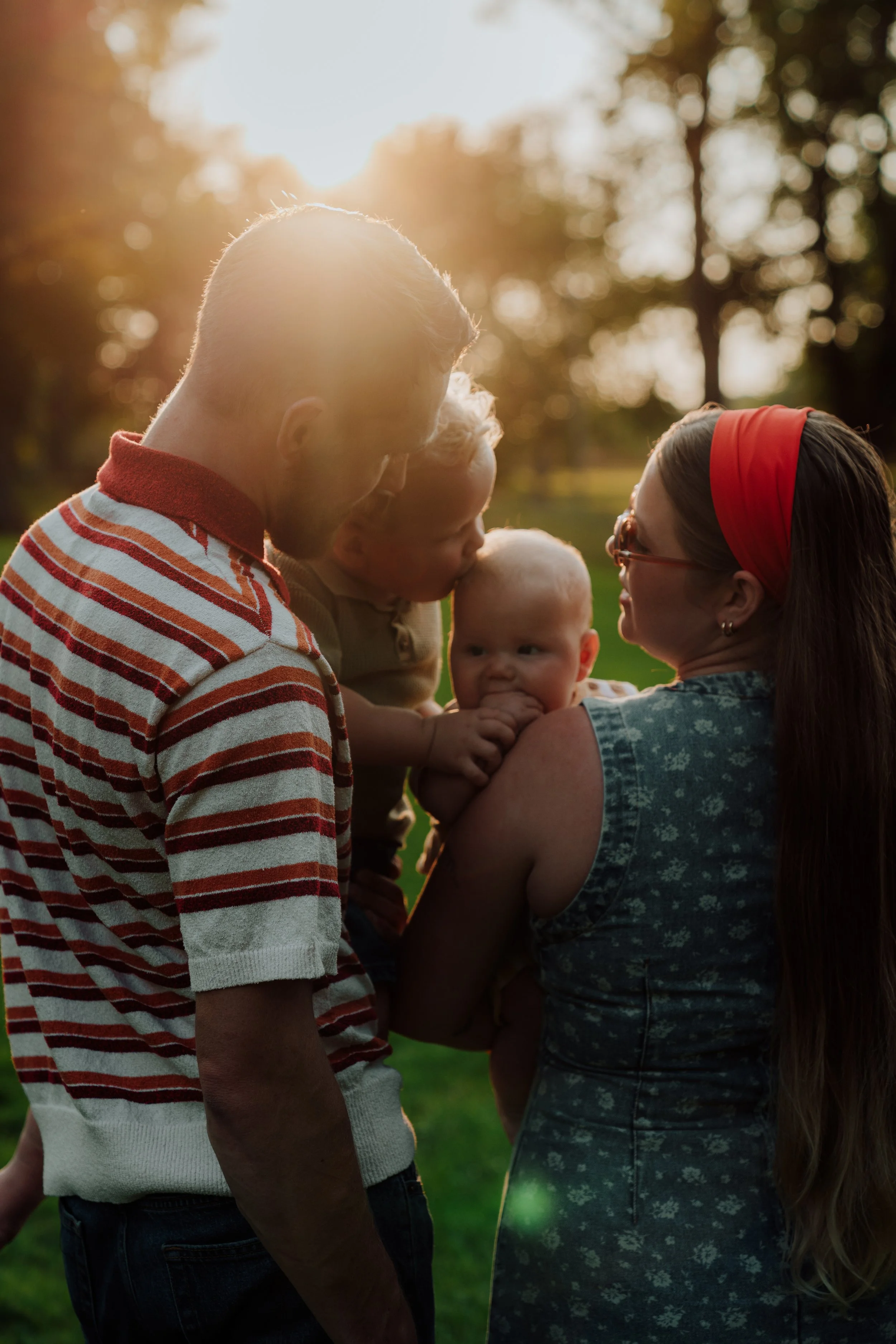 A woman holding a baby with a man and another child standing in a park with trees, backlit by the setting sun.