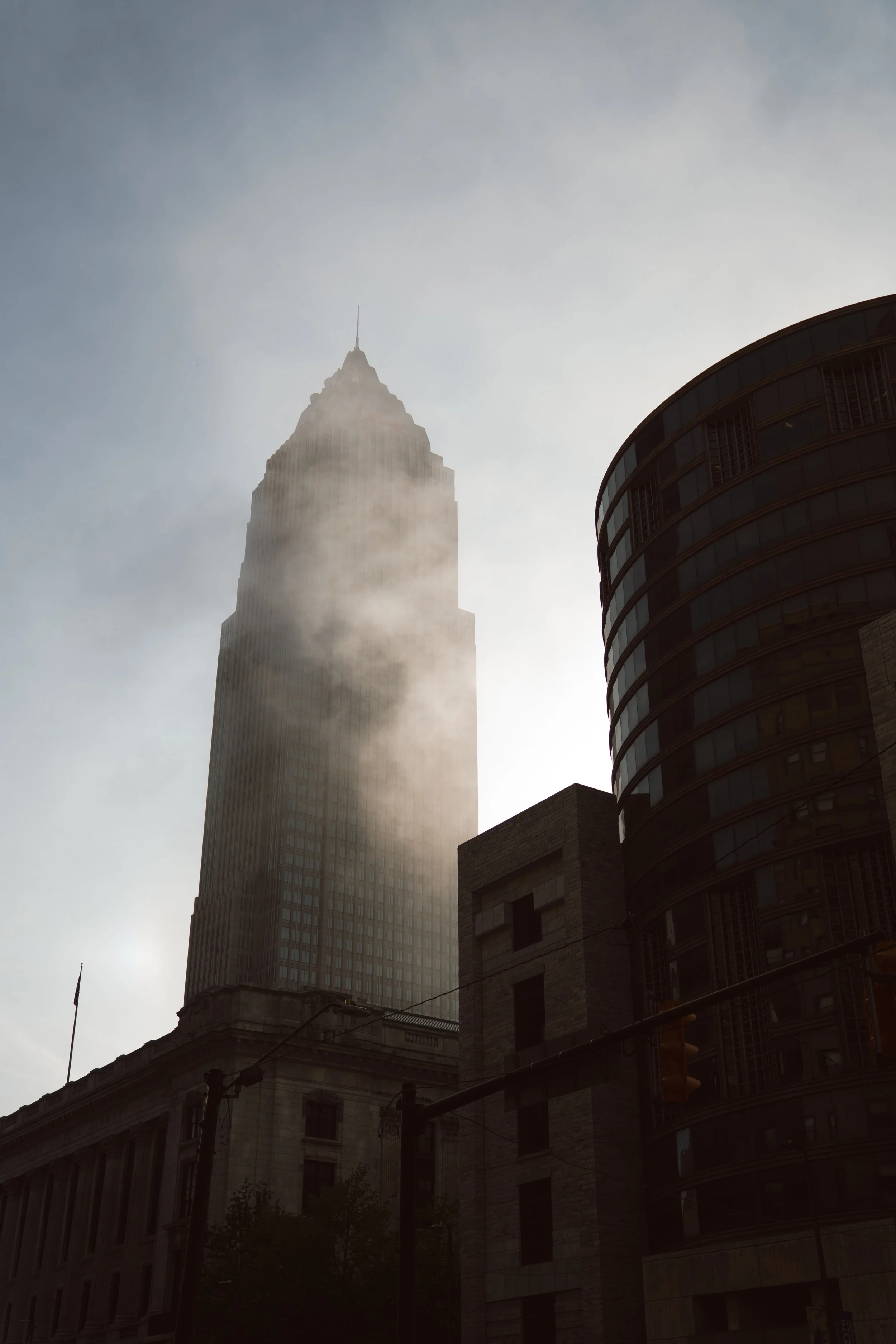 A tall skyscraper with a pointed top partially covered in fog, next to a rounded modern building with glass windows, in an urban cityscape.