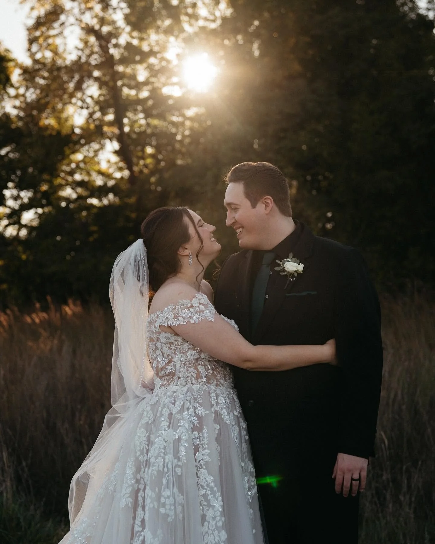 A bride and groom smiling and looking at each other outdoors during sunset, with trees in the background.