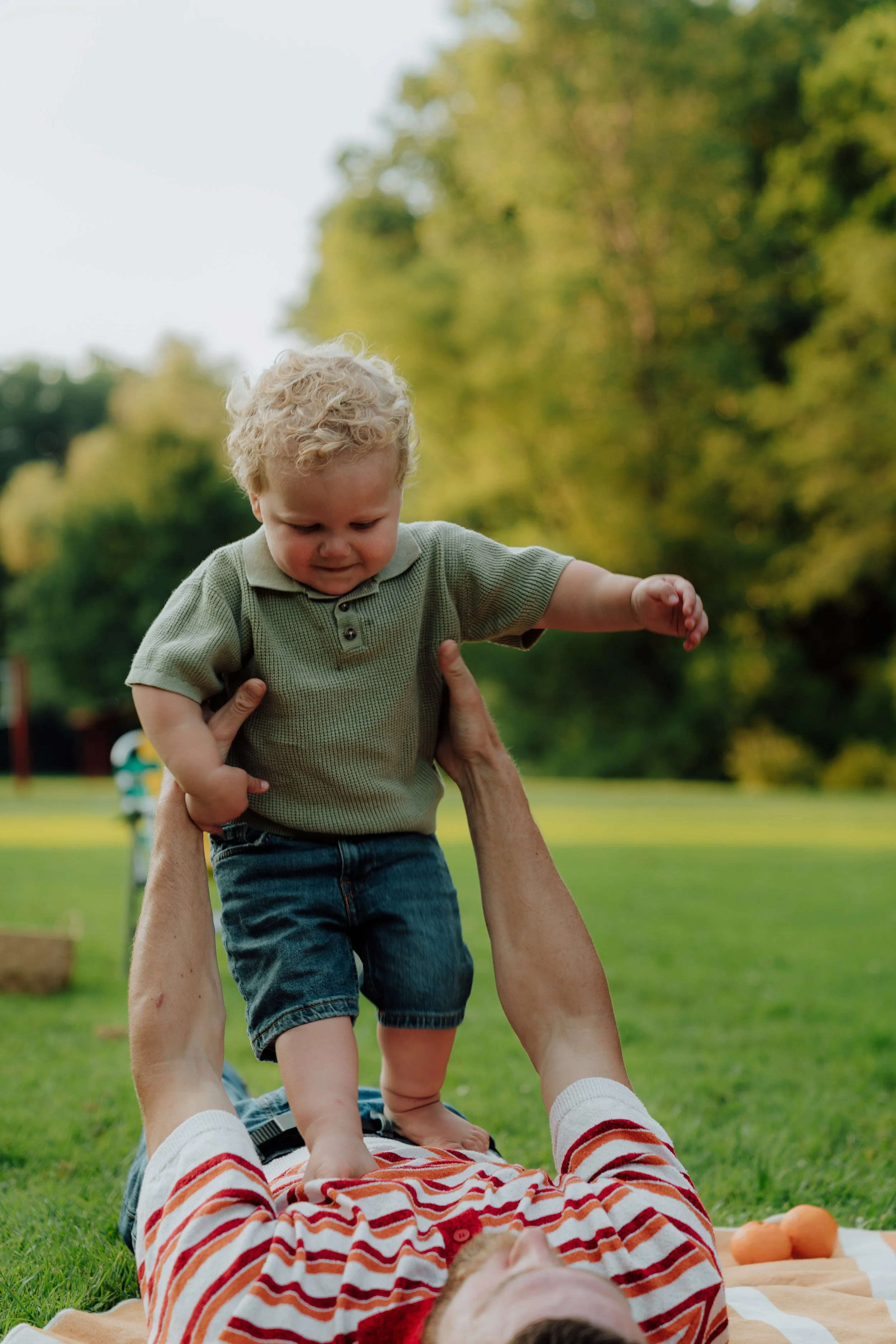 A young boy with curly blonde hair stands on a person's lap outside in a park. The person, lying on the grass, holds the boy's hands. The scene is set on a sunny day with green trees in the background. The person wears a striped orange and white shir
