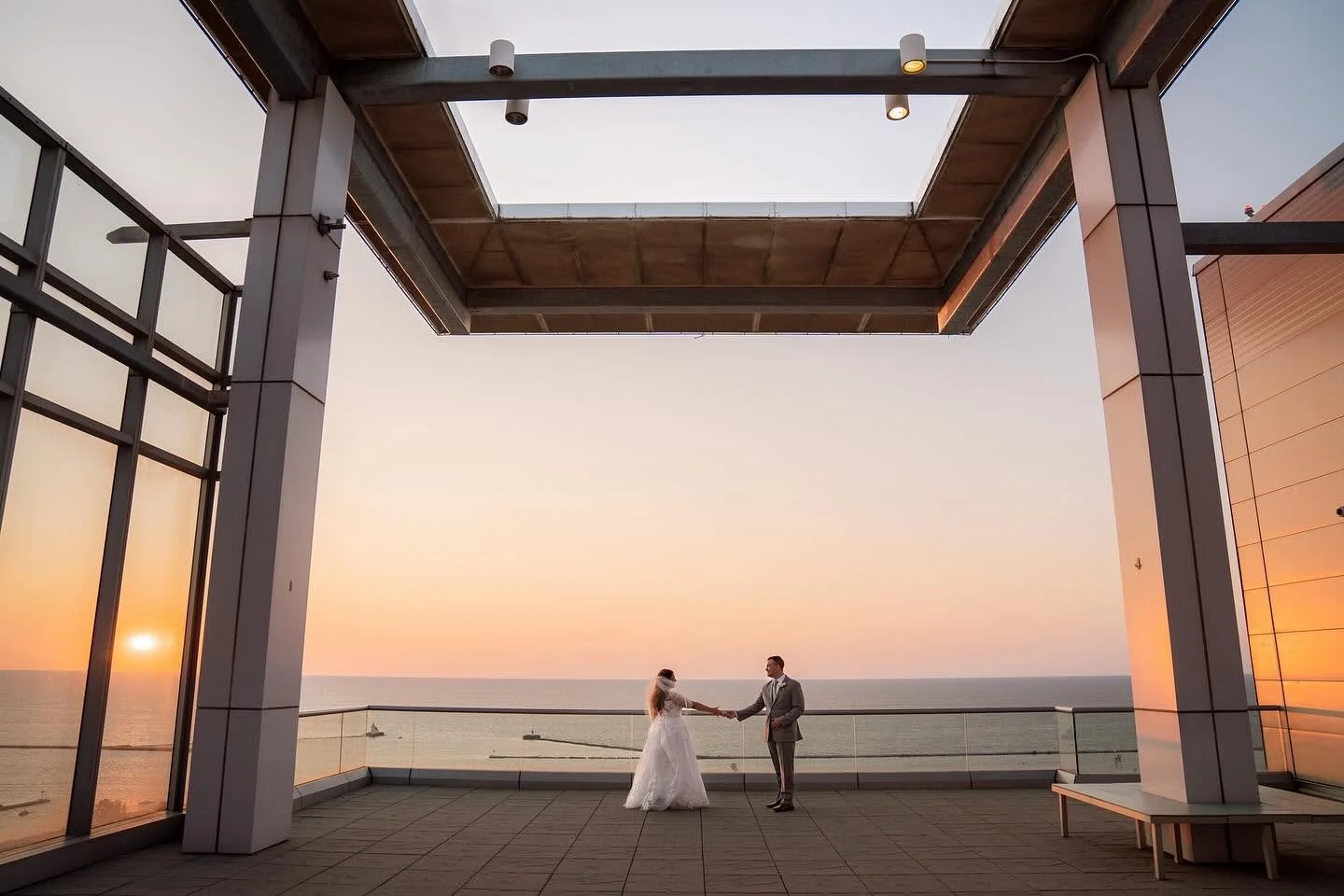 A bride and groom holding hands on a large balcony overlooking the ocean at sunset.