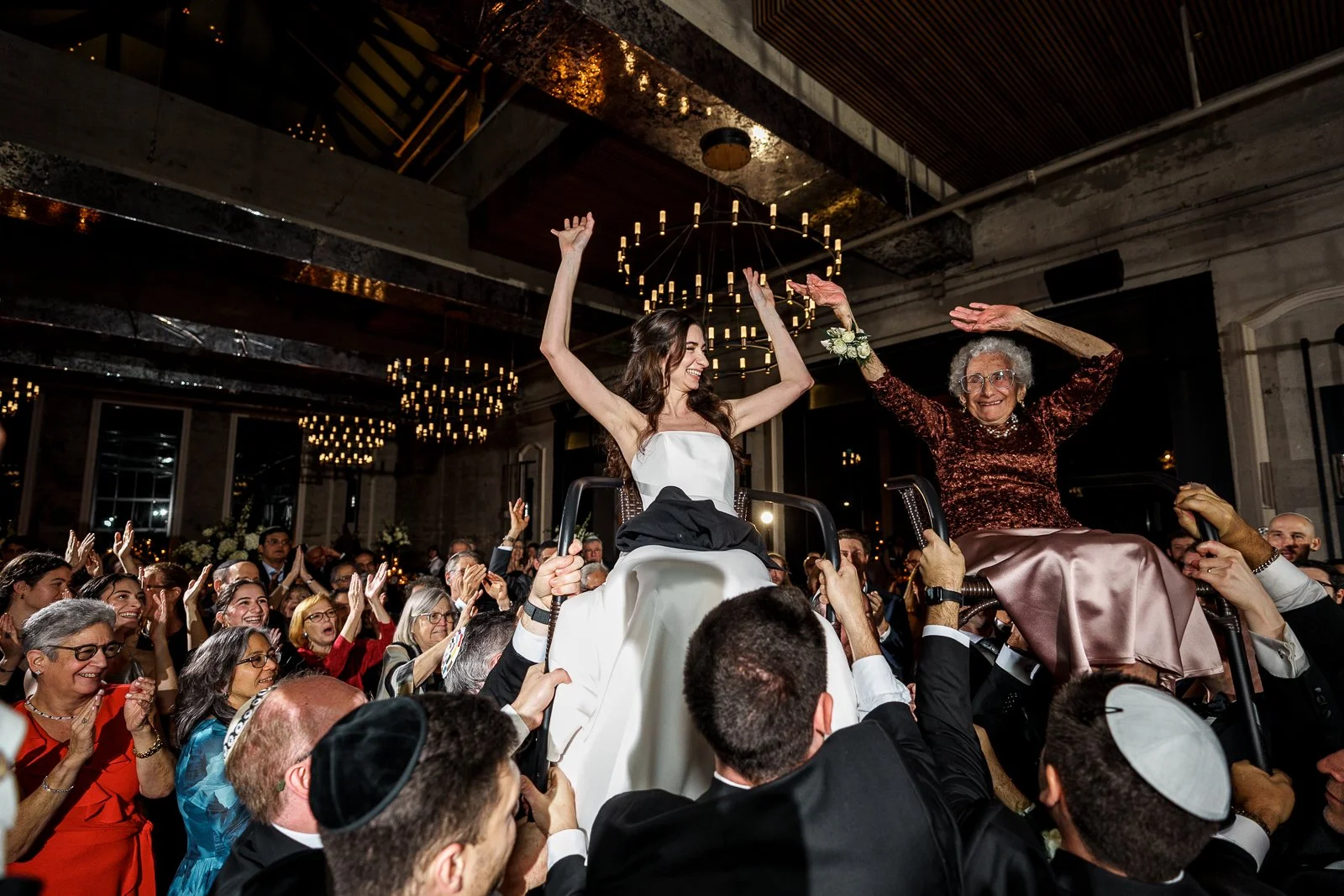 A woman in a wedding dress is lifted in a chair during the hora dance, surrounded by people clapping and celebrating at a wedding reception.