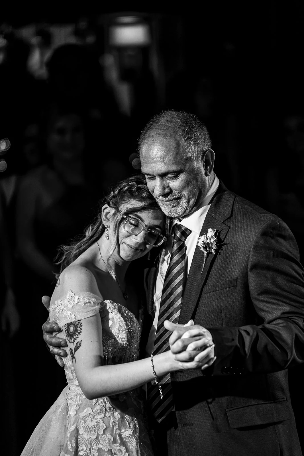 A black and white photo of a young woman and an older man dancing closely together at a wedding. The woman has glasses, a wedding dress with floral embroidery, and a tattoo of a sunflower on her arm. The man is wearing a suit and tie, with a boutonni