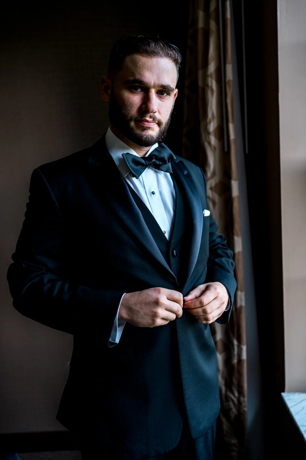 A man in formal black tuxedo with a bow tie, standing near a window with curtains, adjusting his jacket.