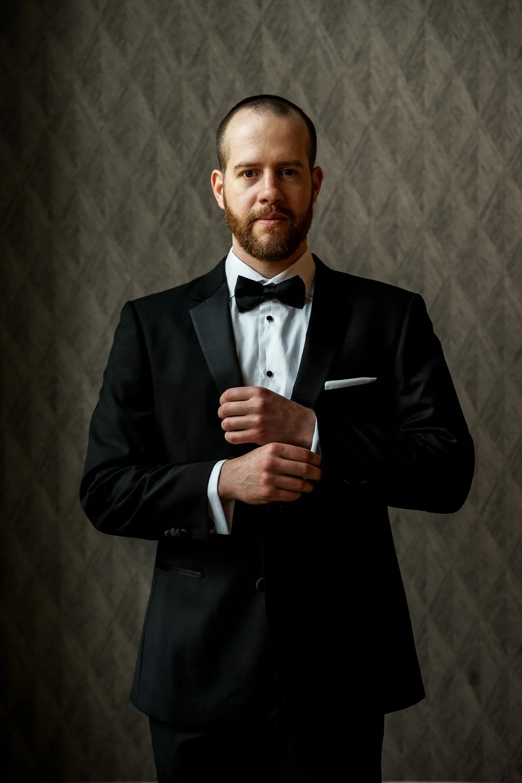 Man in a black tuxedo adjusting his sleeve against a patterned wall.