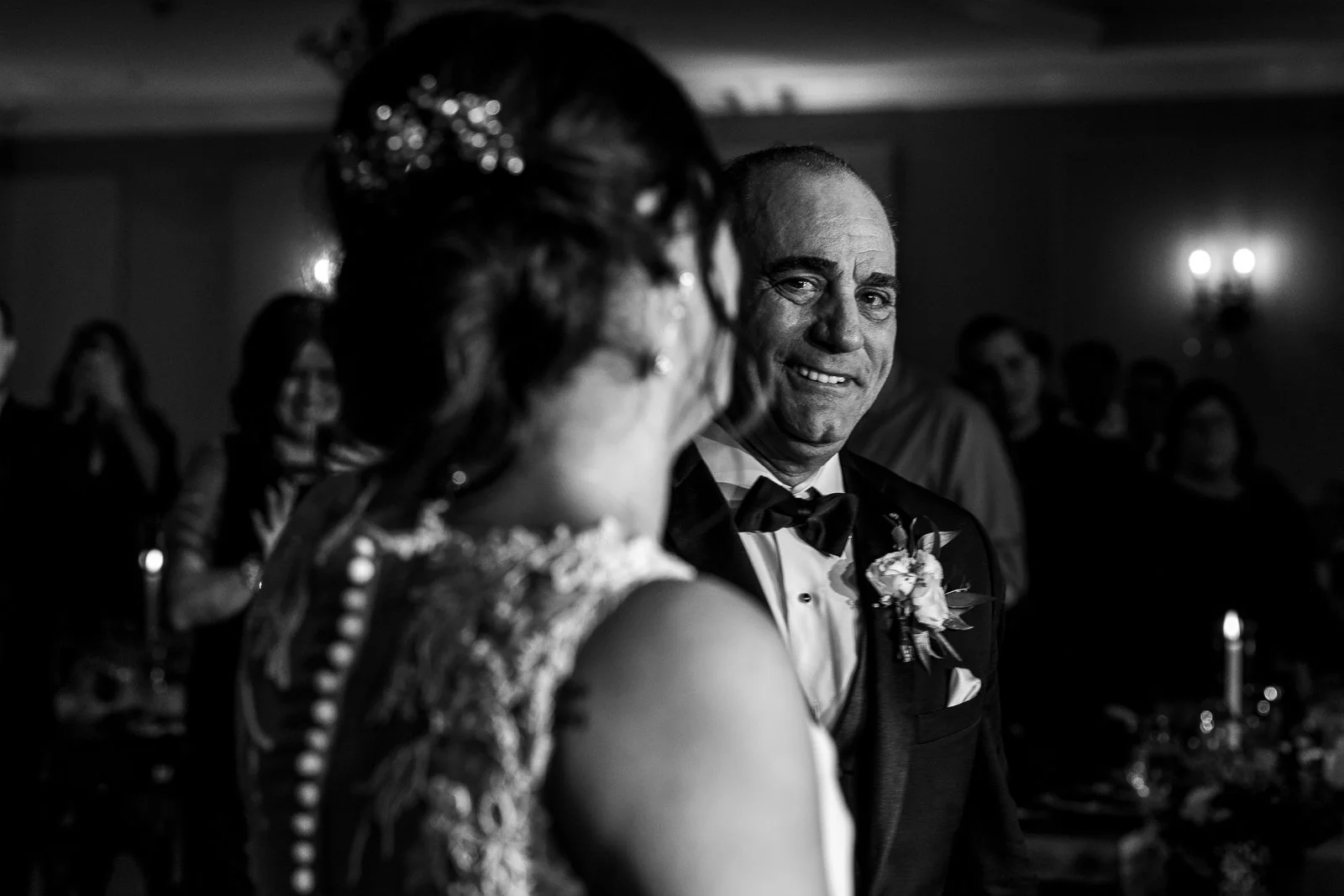 A black and white photo of a man in a tuxedo smiling at a woman in a wedding dress, with guests in the background.