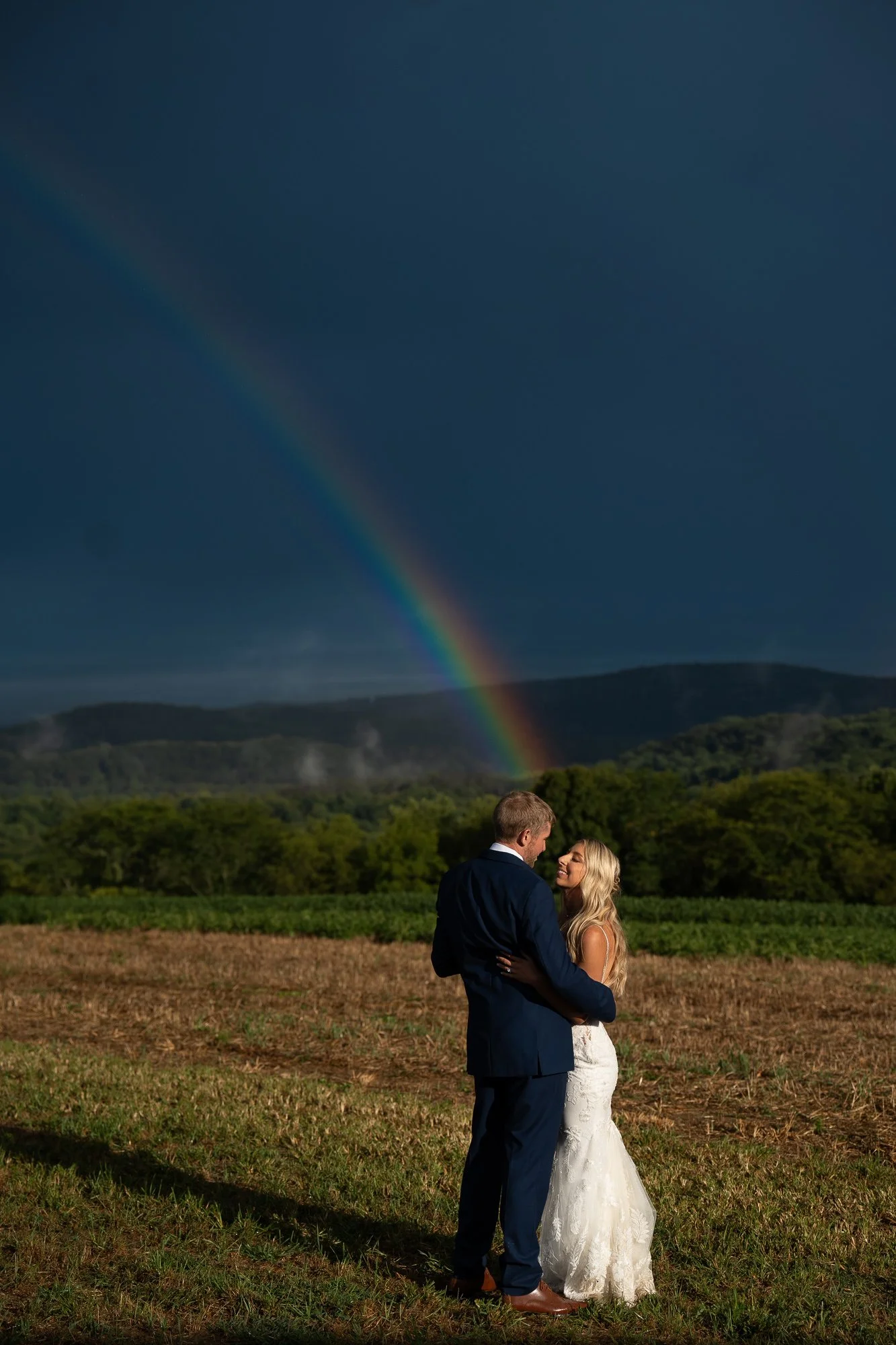 A couple in wedding attire standing outdoors on a grassy field, holding each other and facing each other, with a rainbow and dark stormy sky in the background.