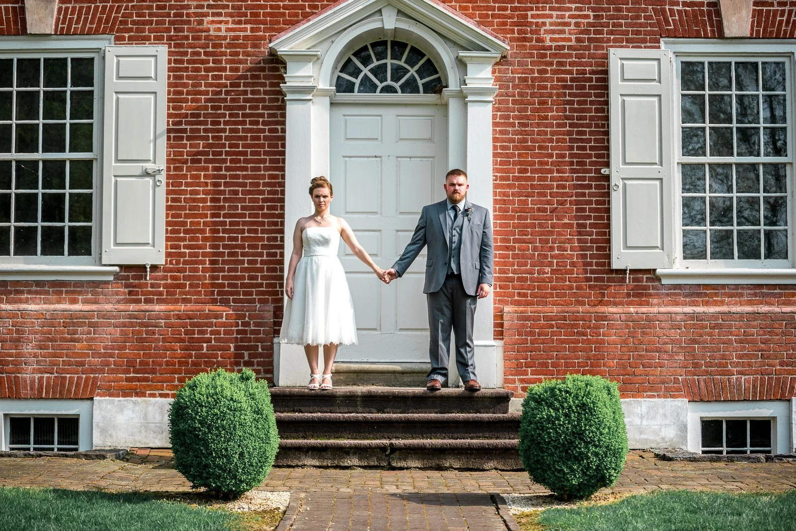 A bride and groom holding hands on the steps of a red brick house with white shutters and a white door, outside during daytime.