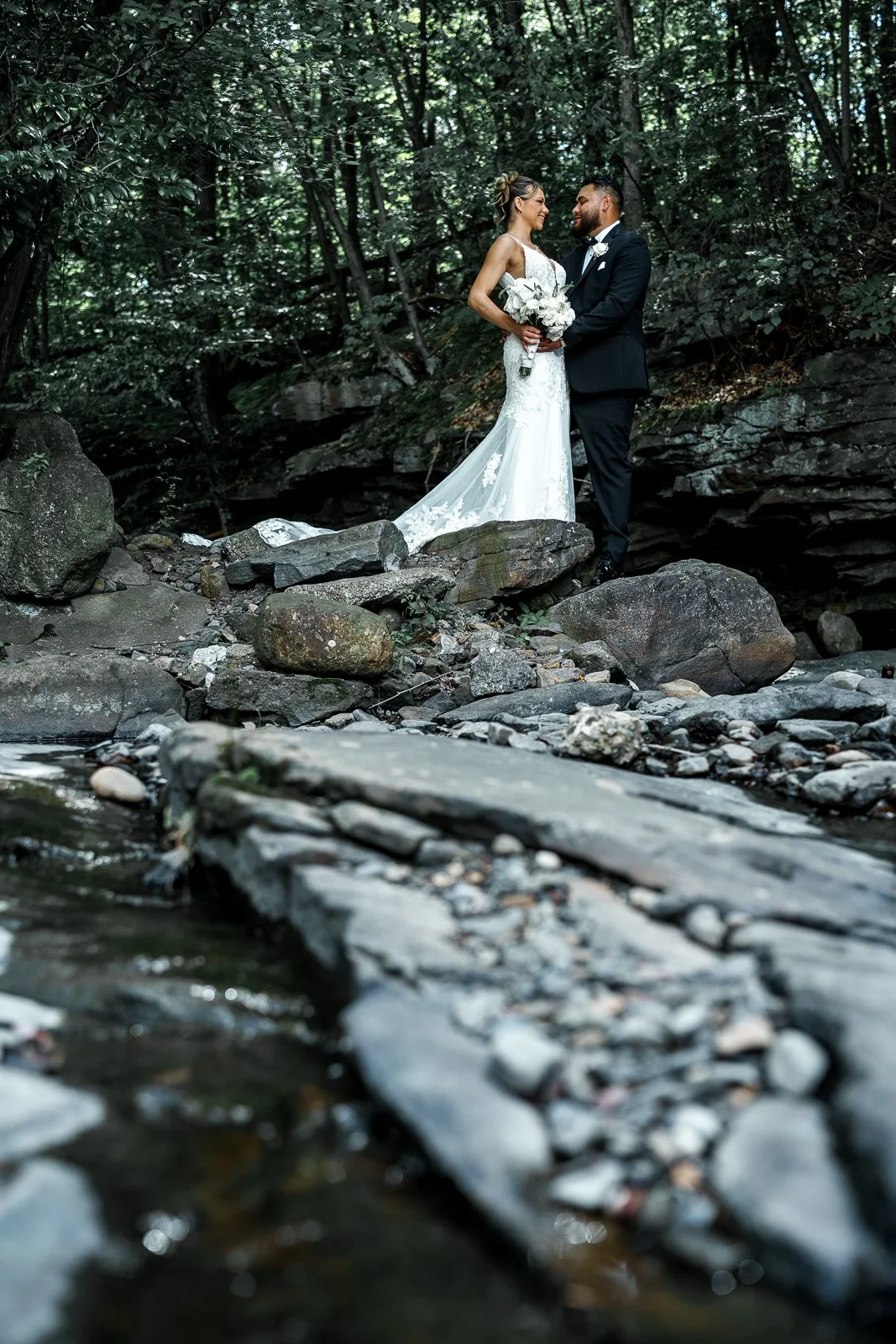 A bride and groom in wedding attire standing on rocks near a creek in a forest, facing each other and smiling.