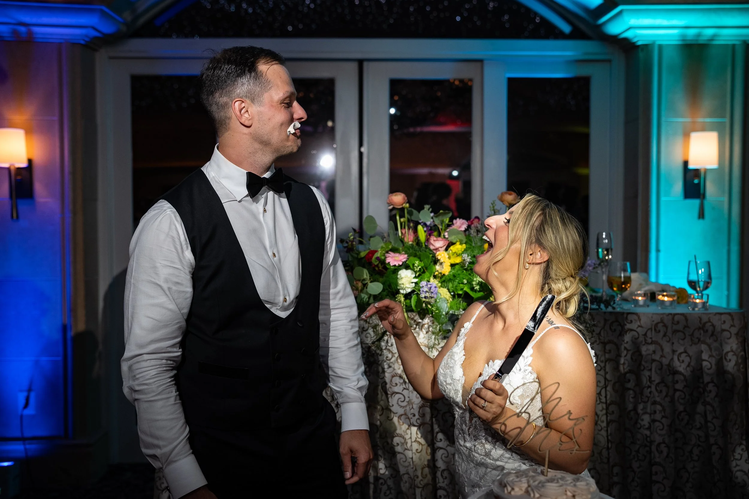 A bride and groom at a wedding reception, with the bride holding a cake knife, laughing and smiling at each other, cake and flowers on the table behind them.