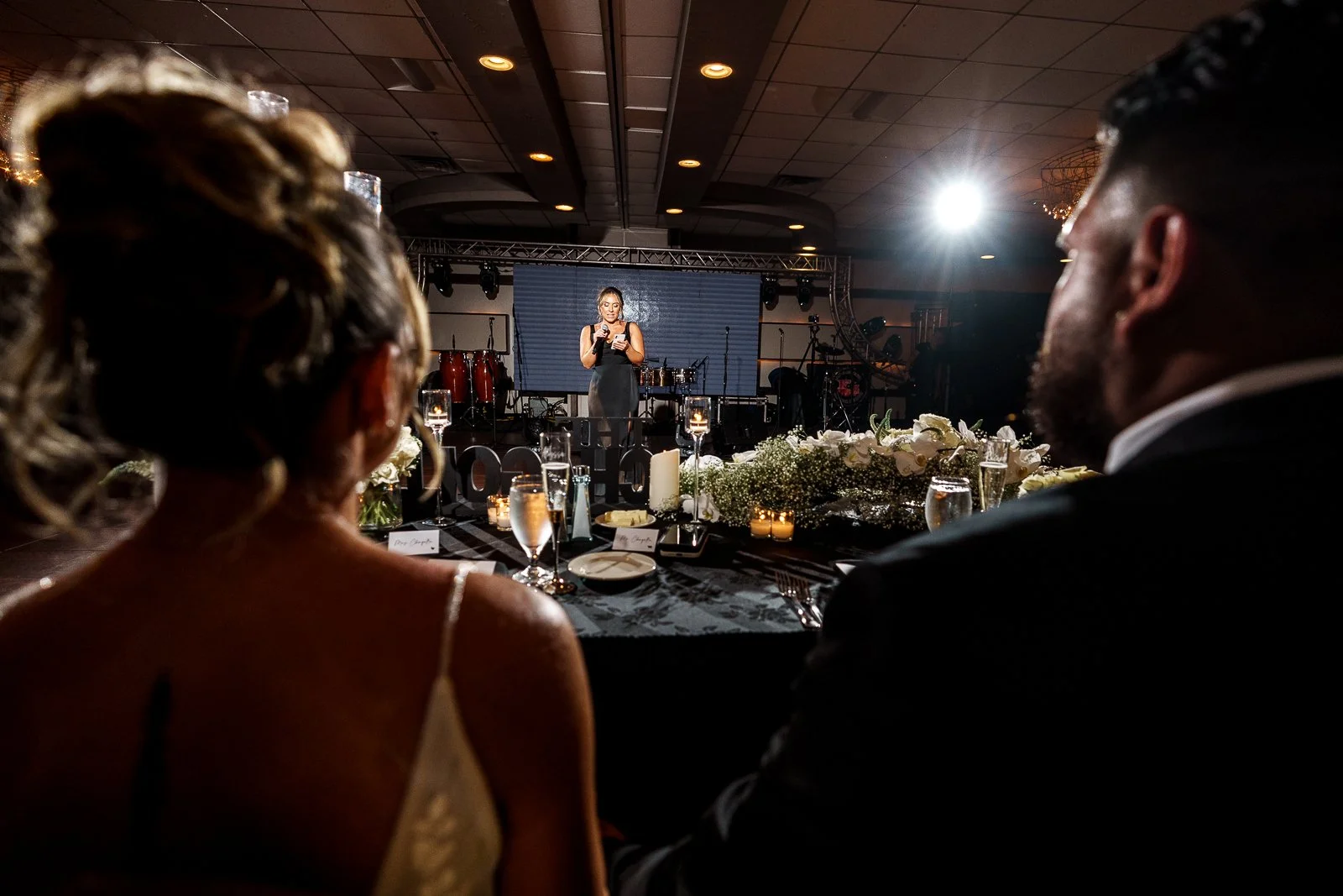 A woman in a black dress performing on stage at a formal event. Audience members are seated at a decorated table with flowers, candles, wine glasses, and plates, watching her.