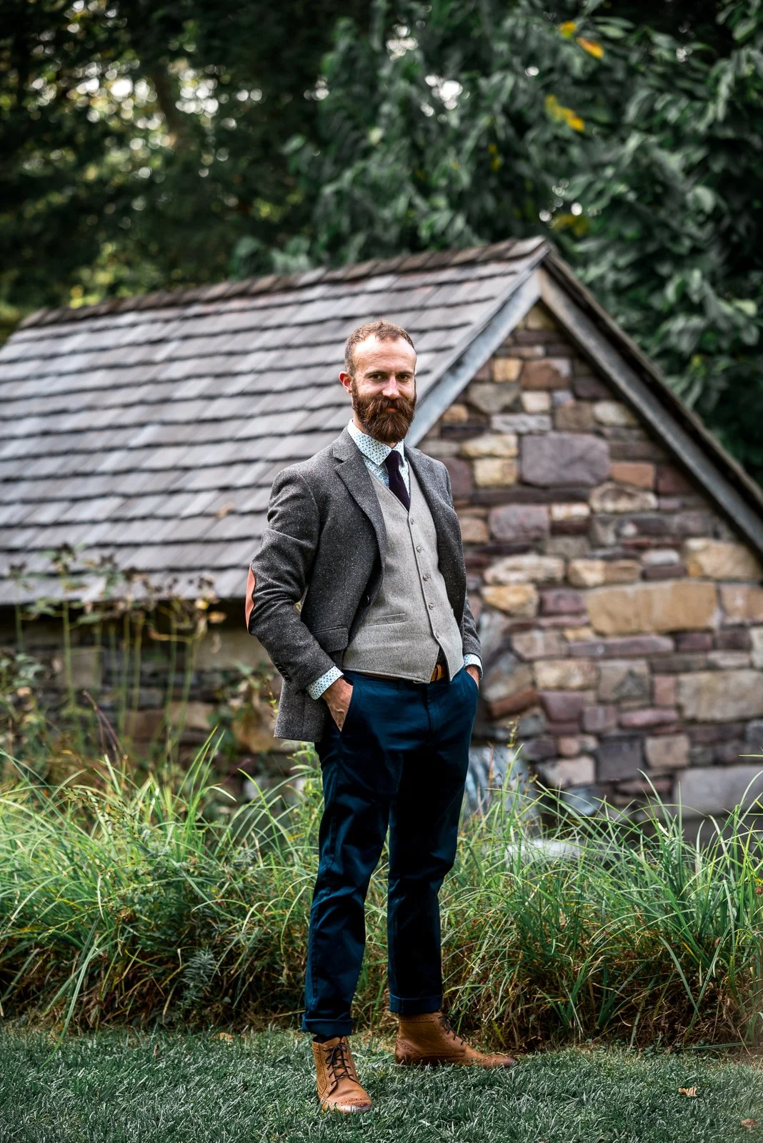 A man with a beard wearing a gray blazer, vest, dress shirt, dark pants, and tan boots standing outdoors in front of a stone cottage with lush green trees in the background.
