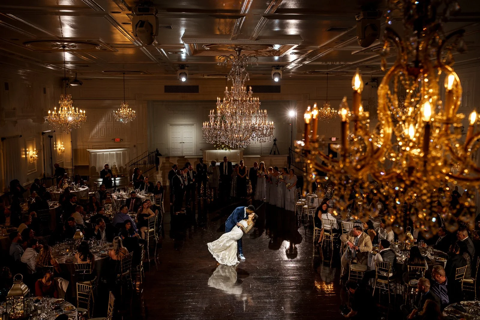 A bride and groom dancing at their wedding reception in an elegant banquet hall with chandeliers, surrounded by guests seated at decorated tables.