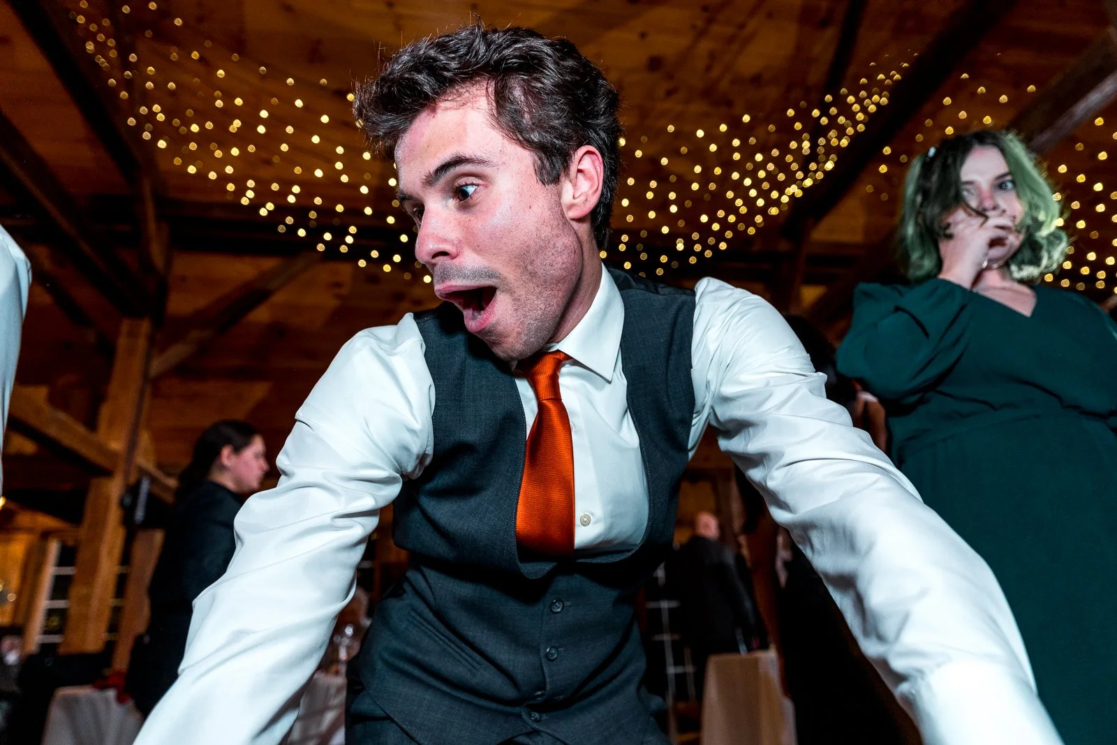 A man in a white shirt, gray vest, and orange tie appears shocked or surprised at a social event held in a wooden venue decorated with string lights.