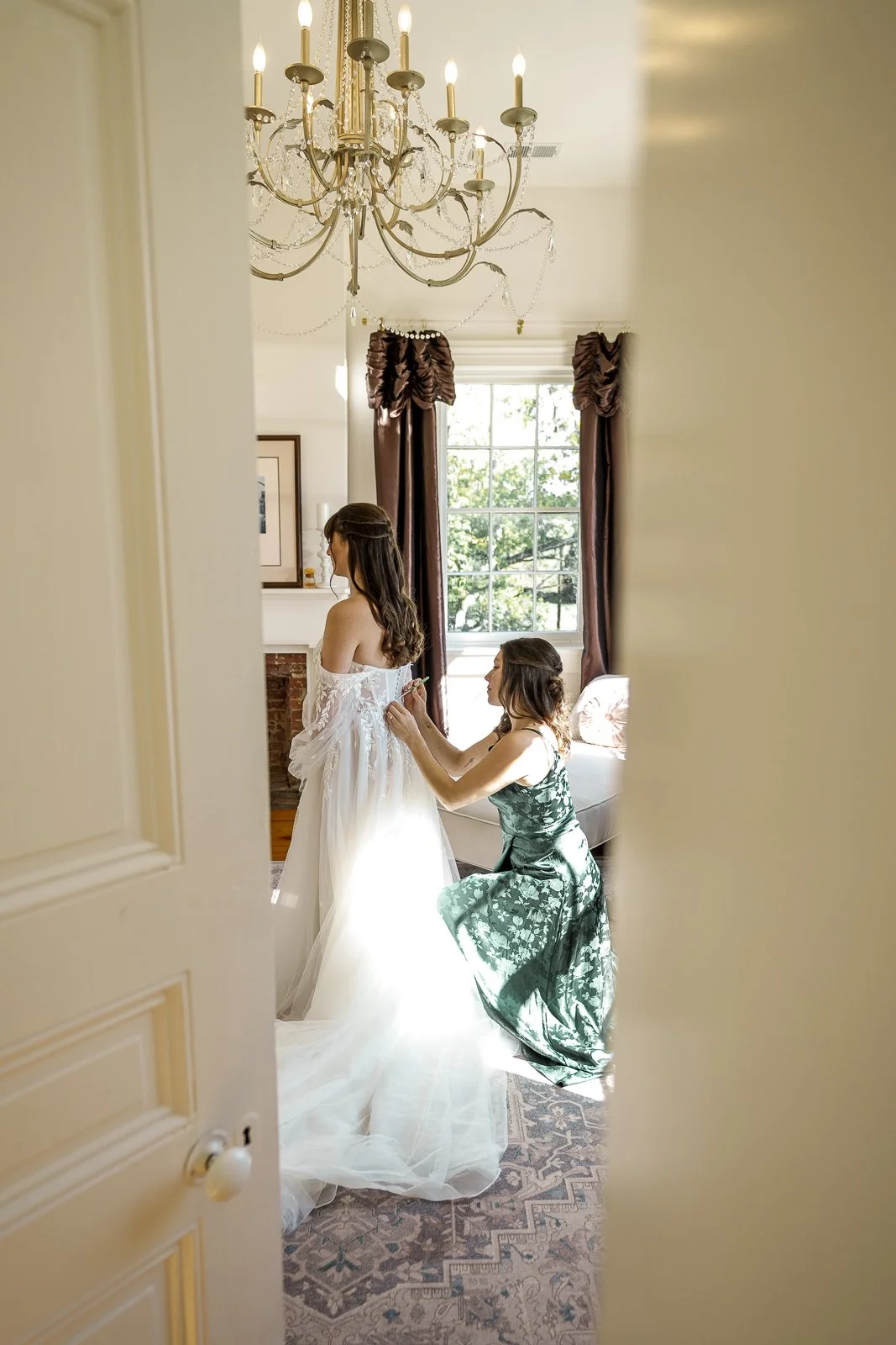 A woman helps a bride with her wedding dress in a well-lit room with a chandelier, large window, and elegant curtains.