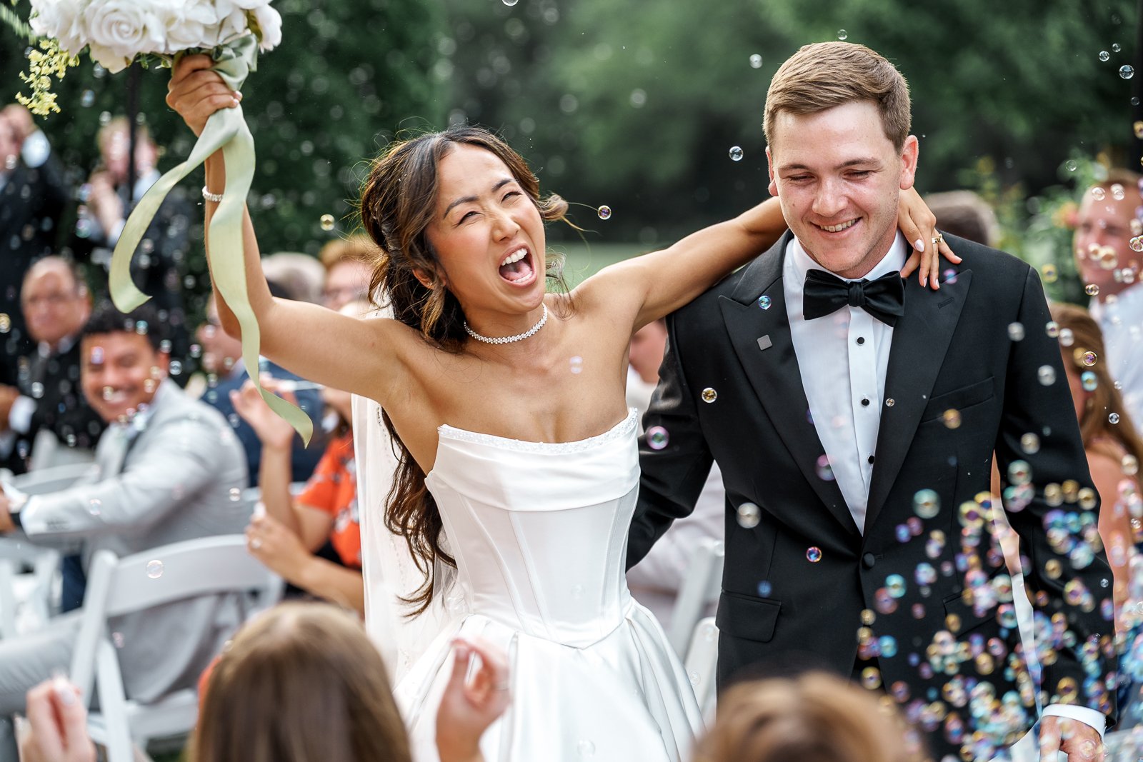 Bride and groom celebrating at their outdoor wedding reception, with the bride cheering and holding a bouquet, guests in the background, and confetti falling.