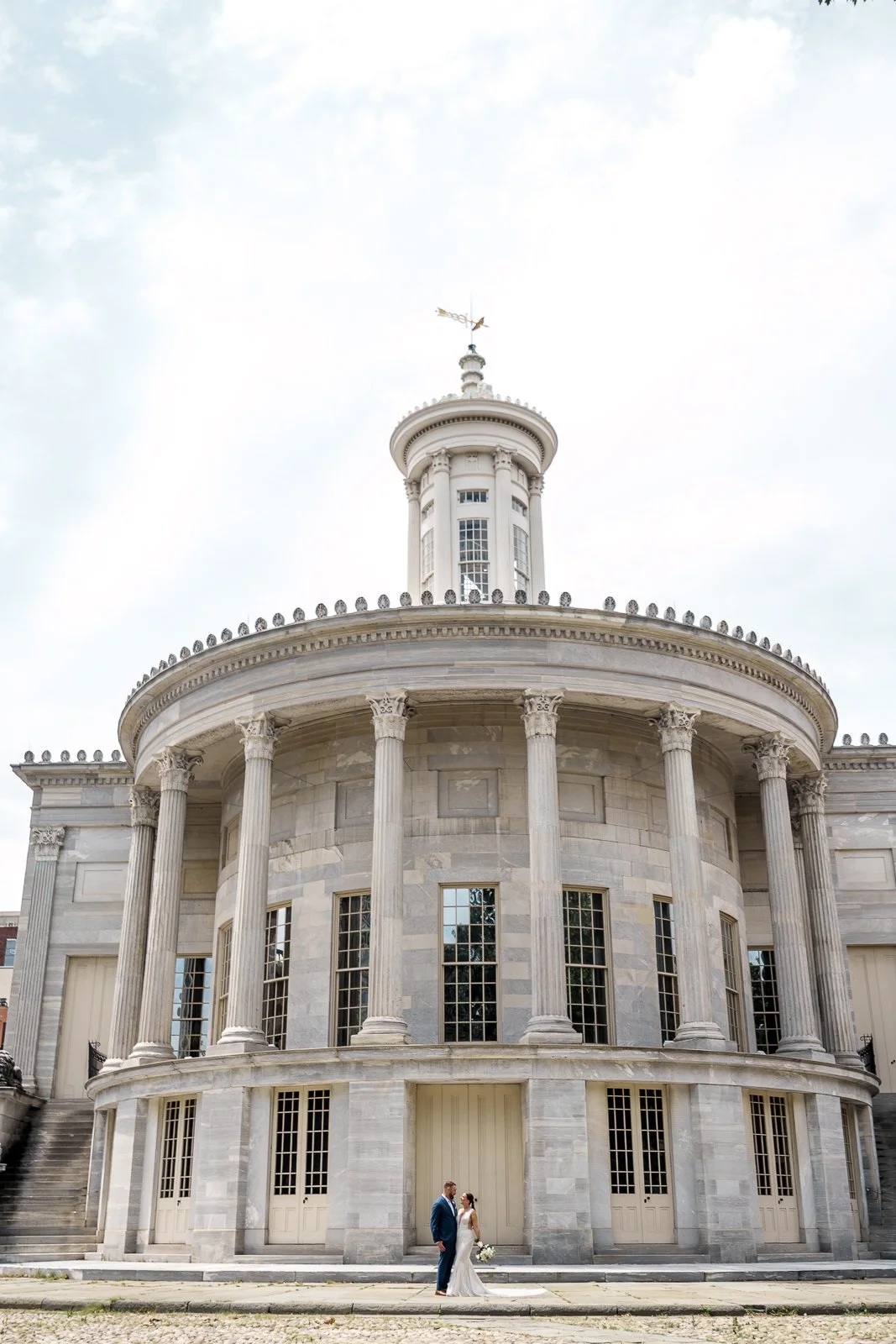 A bride and groom standing in front of a historic building with large columns and a round balcony, with a cloudy sky above.