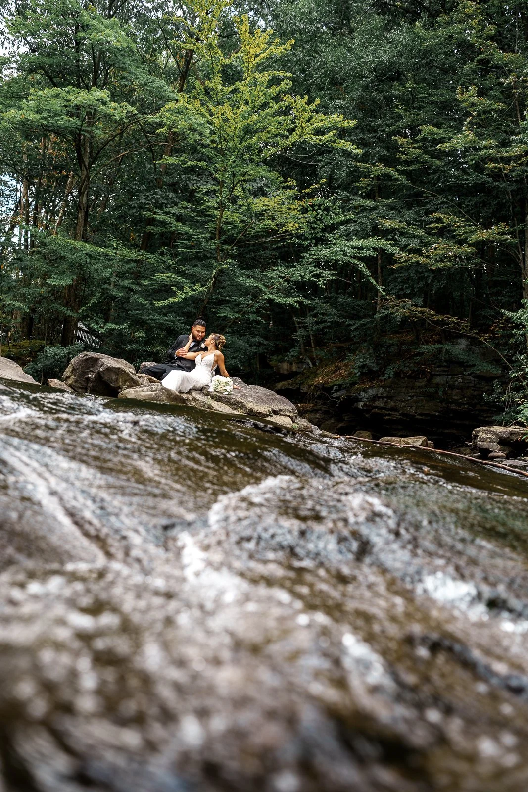 A wedding couple sitting on rocks by a river, surrounded by dense green trees.